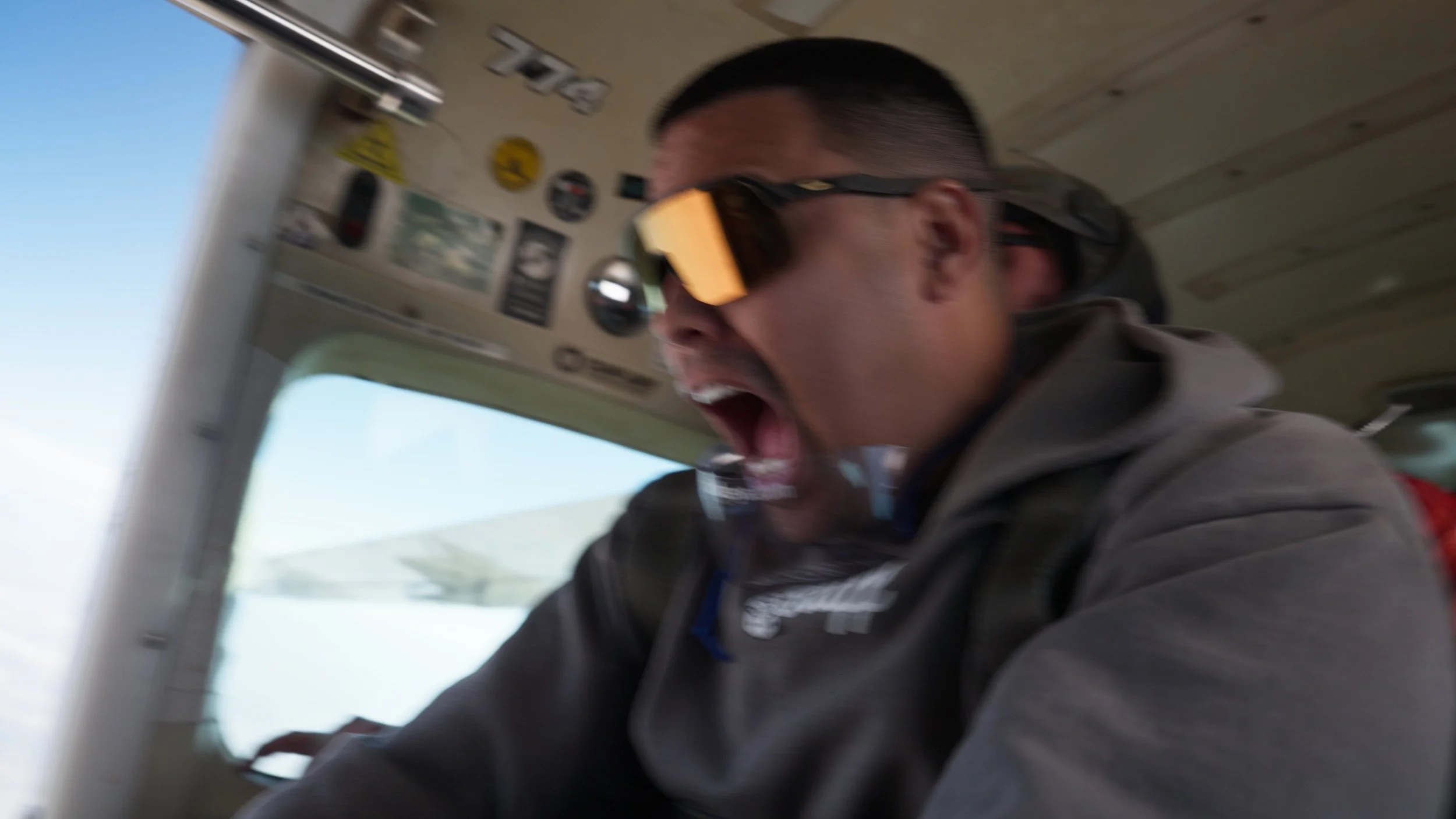 A person wearing sunglasses with orange lenses and a gray jacket, inside the cockpit of an aircraft, appears to be shouting or expressing excitement.