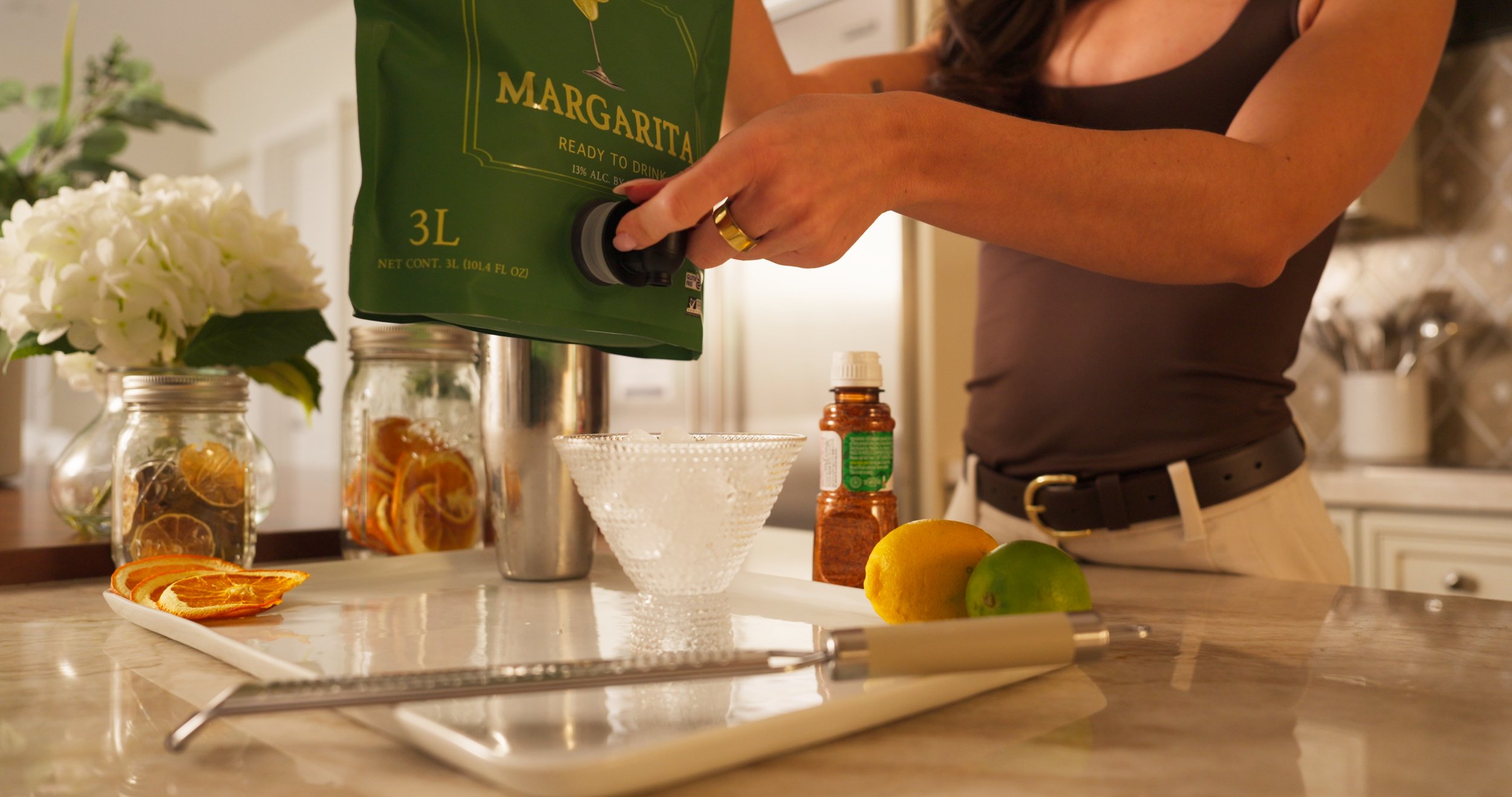 A person pours margarita mix from a green 3-liter bag into a glass on a kitchen countertop. There are sliced oranges, a lemon, and a lime on the counter, along with a grater, a spice jar, and a decorative flower arrangement.
