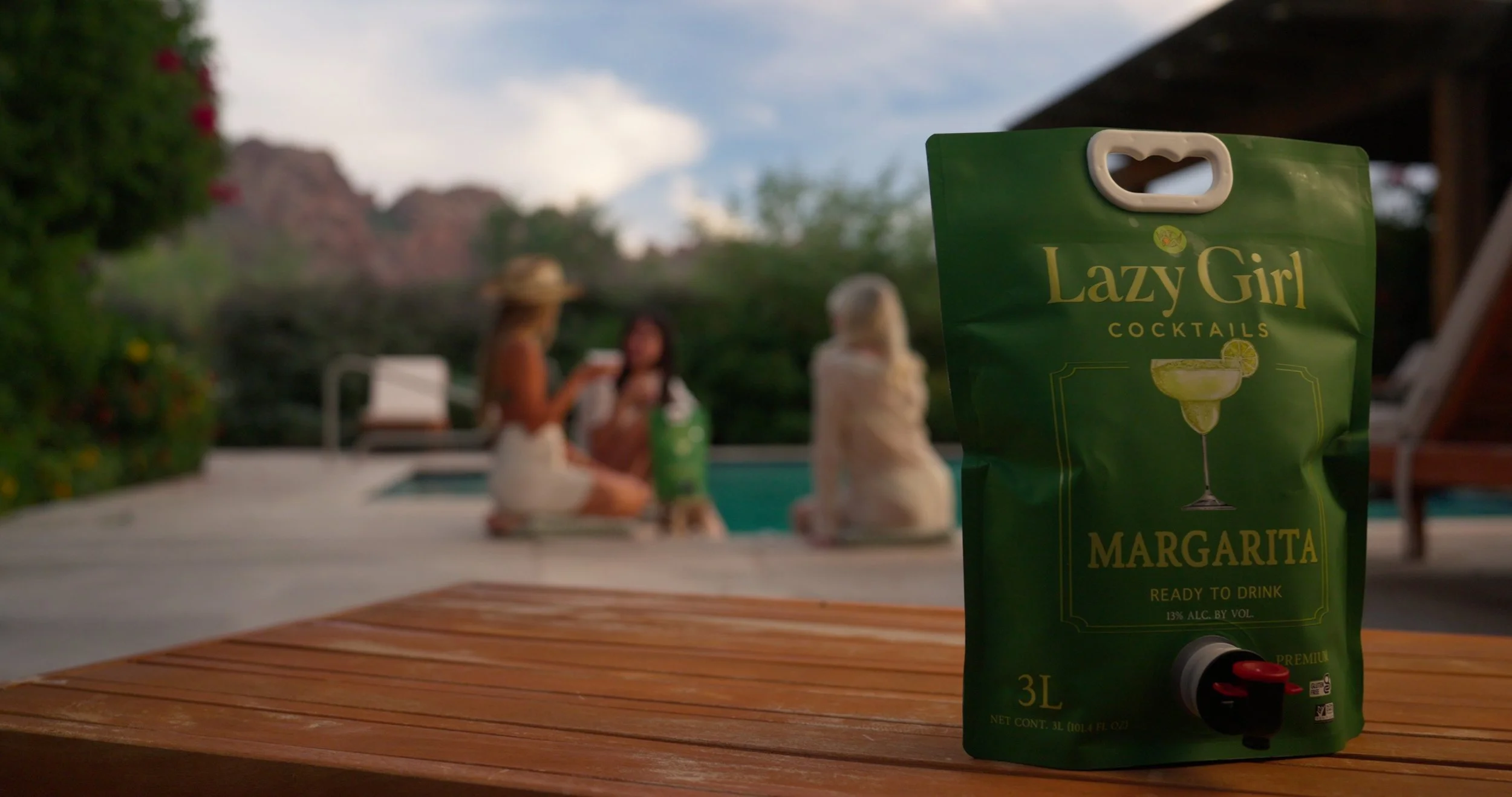 A green bag labeled 'Lazy Girl Cocktails Margarita' on a wooden surface with a blurred background of three women sitting by a pool enjoying drinks.