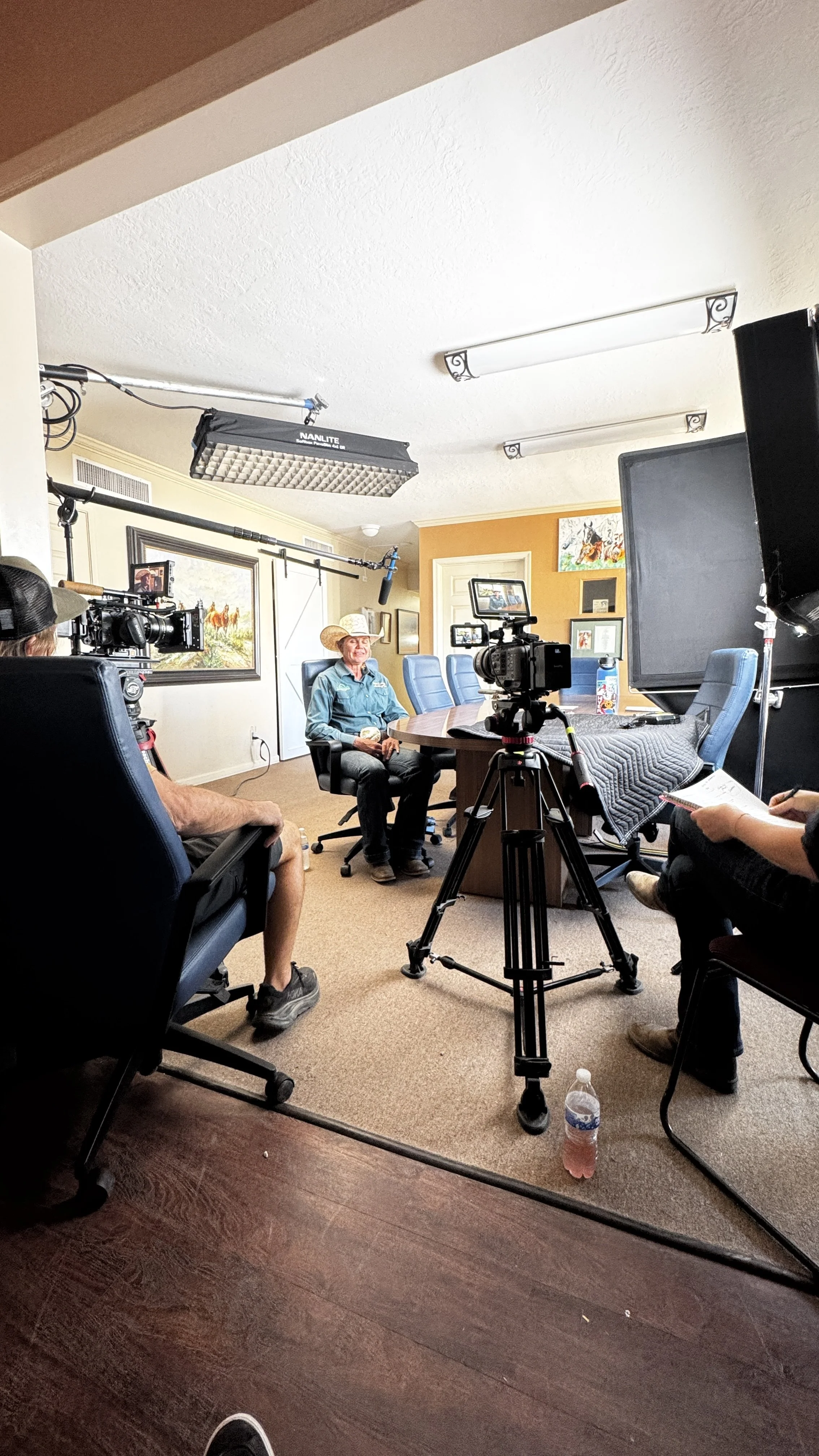 Inside a filming studio, a woman wearing a cowboy hat sits at a conference table being interviewed, with cameras and lighting equipment around her.