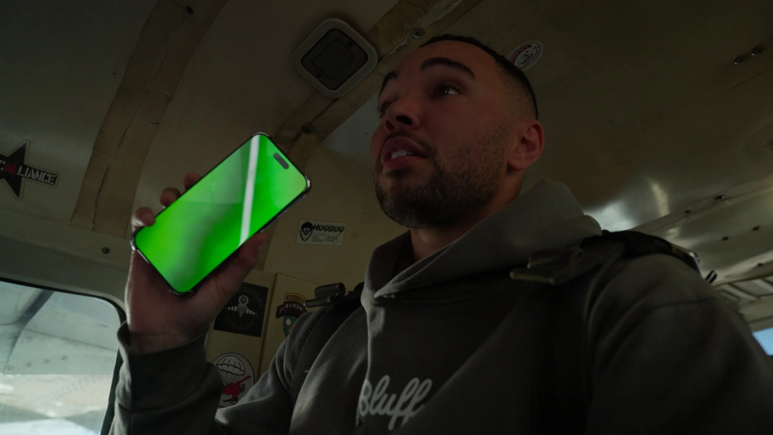 A man with a beard and a short haircut is holding a smartphone with a bright green screen close to his face inside a vehicle, possibly a bus or a plane. The background shows posters on the wall.