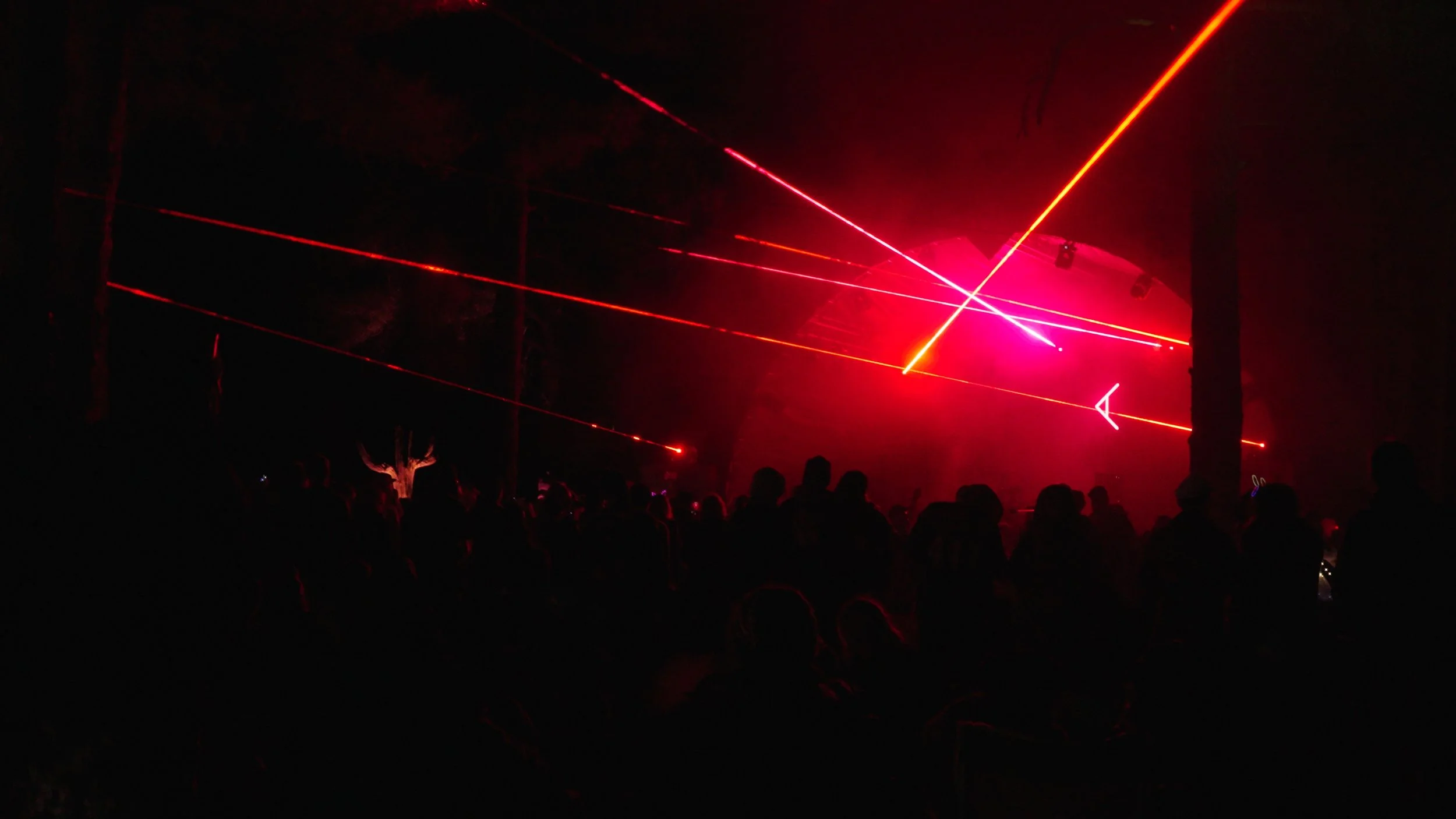 Dark scene with silhouettes of people watching a laser light show with red and pink lasers in the night sky.