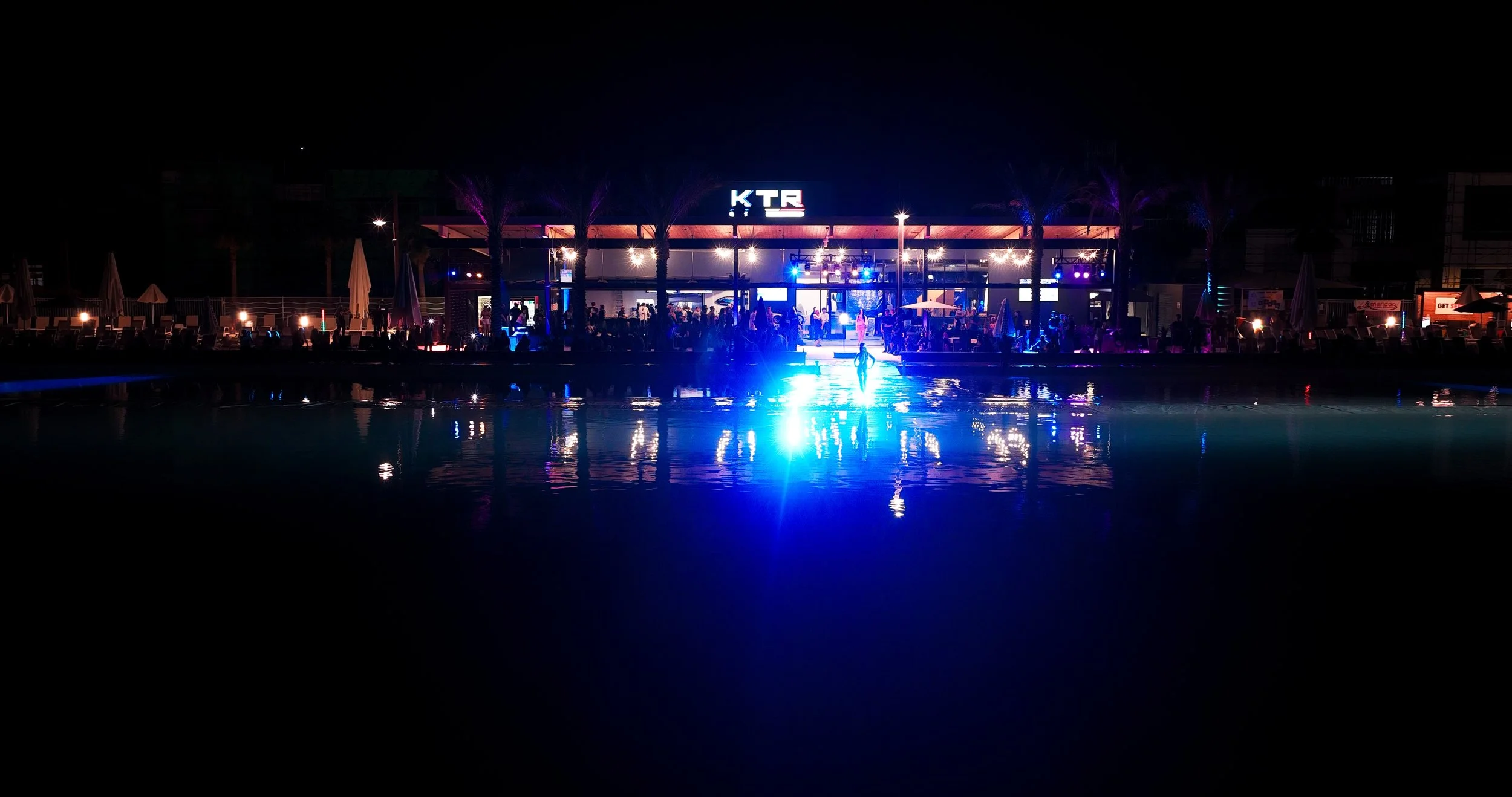 Nighttime scene at a poolside bar with colorful lighting, people socializing, and a person walking on the reflecting pool surface.