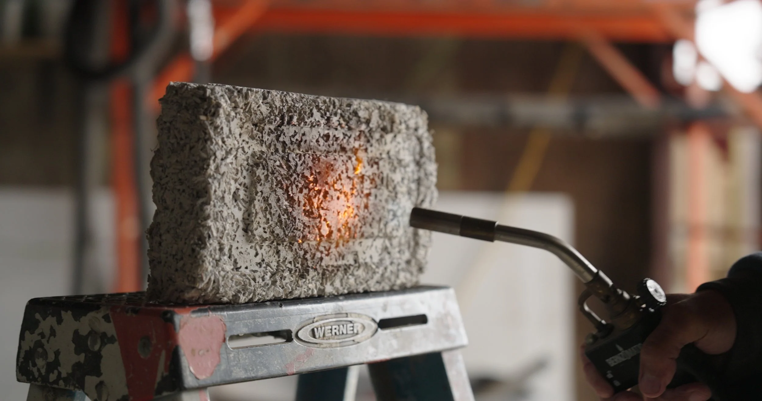 A worker is welding a rough-textured concrete block on a metal workbench in a workshop, with sparks and bright light from the welding process.