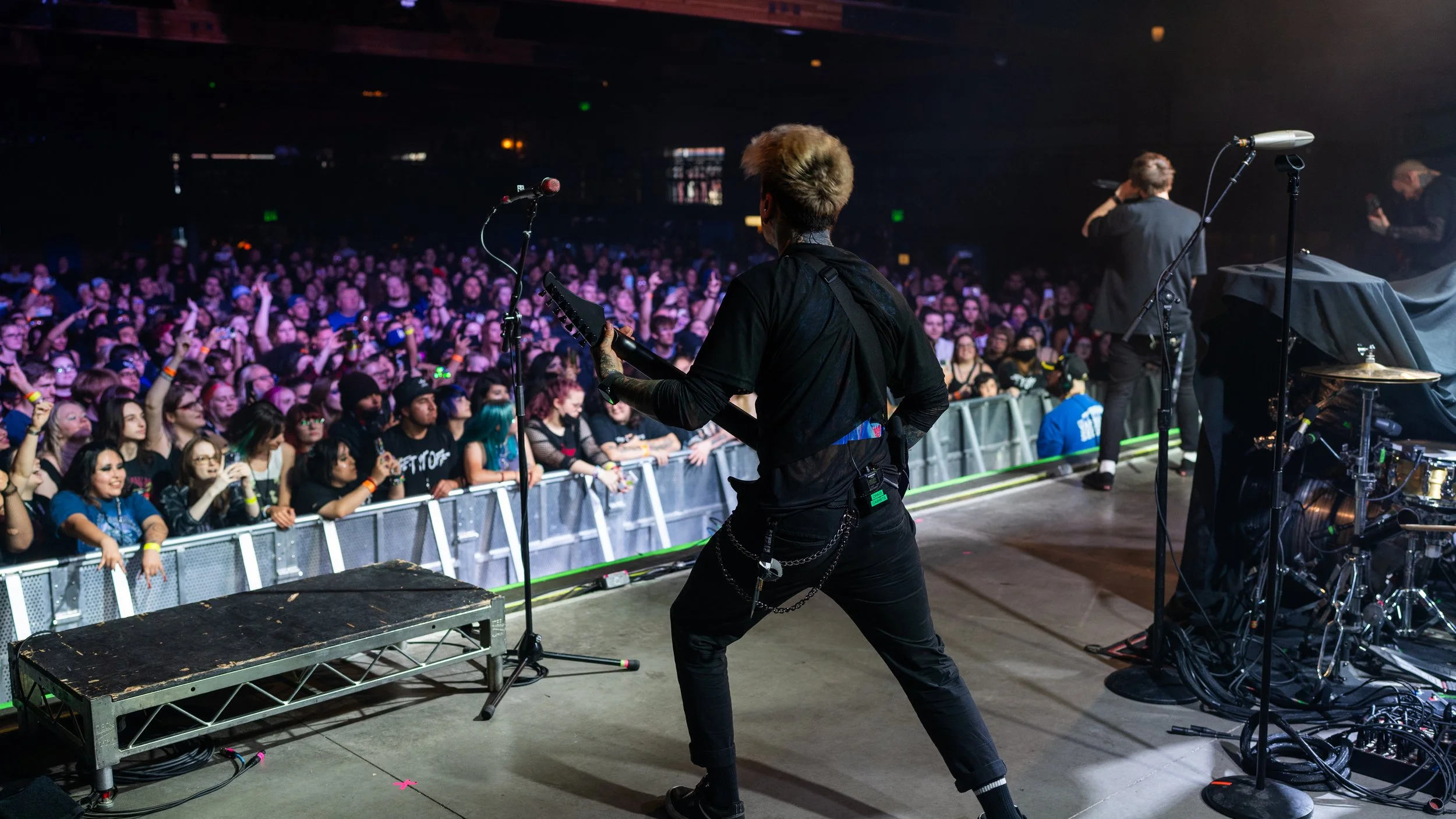 A musician playing guitar on stage during a concert, facing an audience with many people, some taking photos and videos, in a dark concert hall.