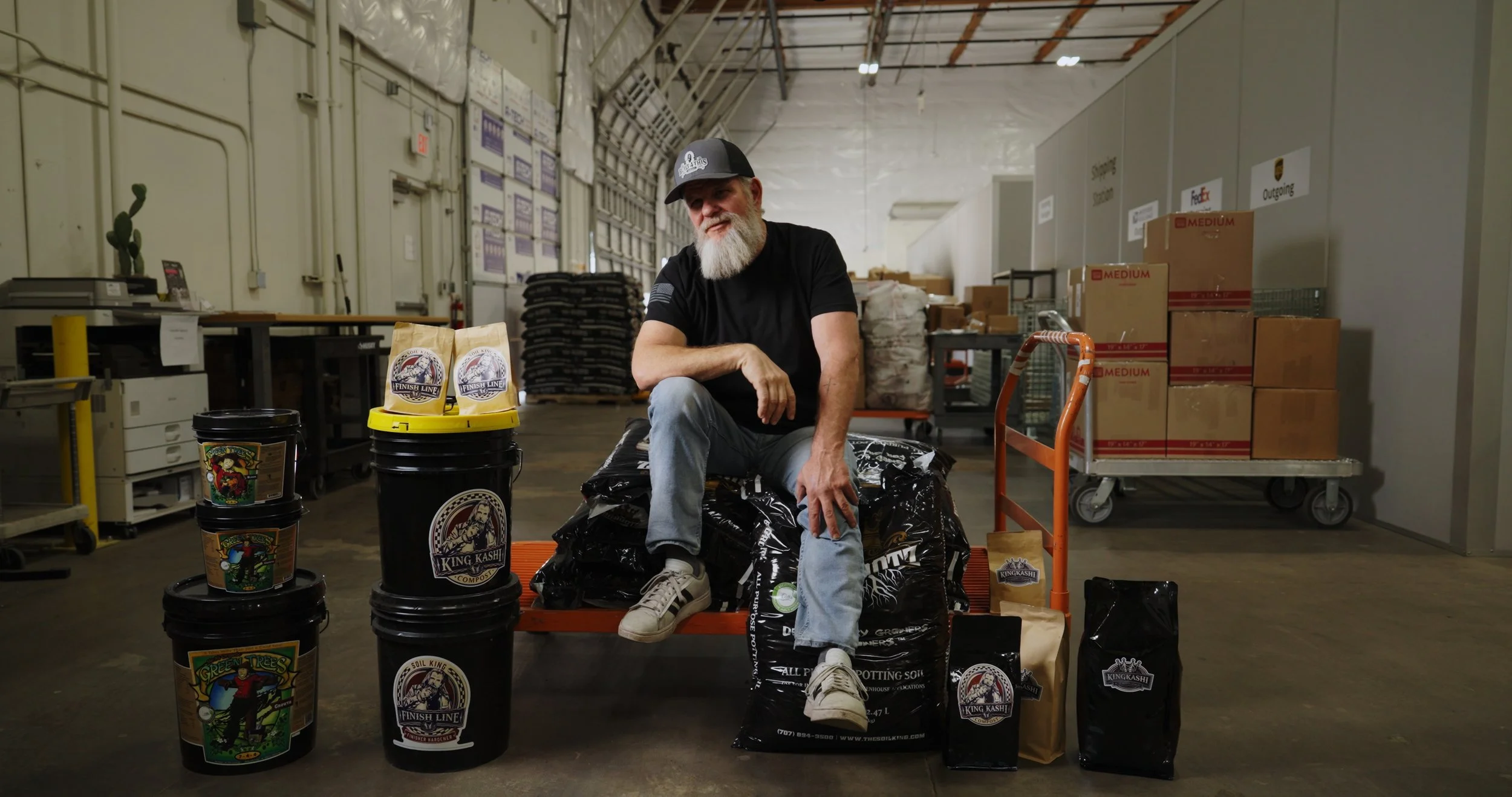 A man with a gray beard and black cap sitting on an orange cart in a warehouse surrounded by bags and containers of potting soil and plant fertilizer.