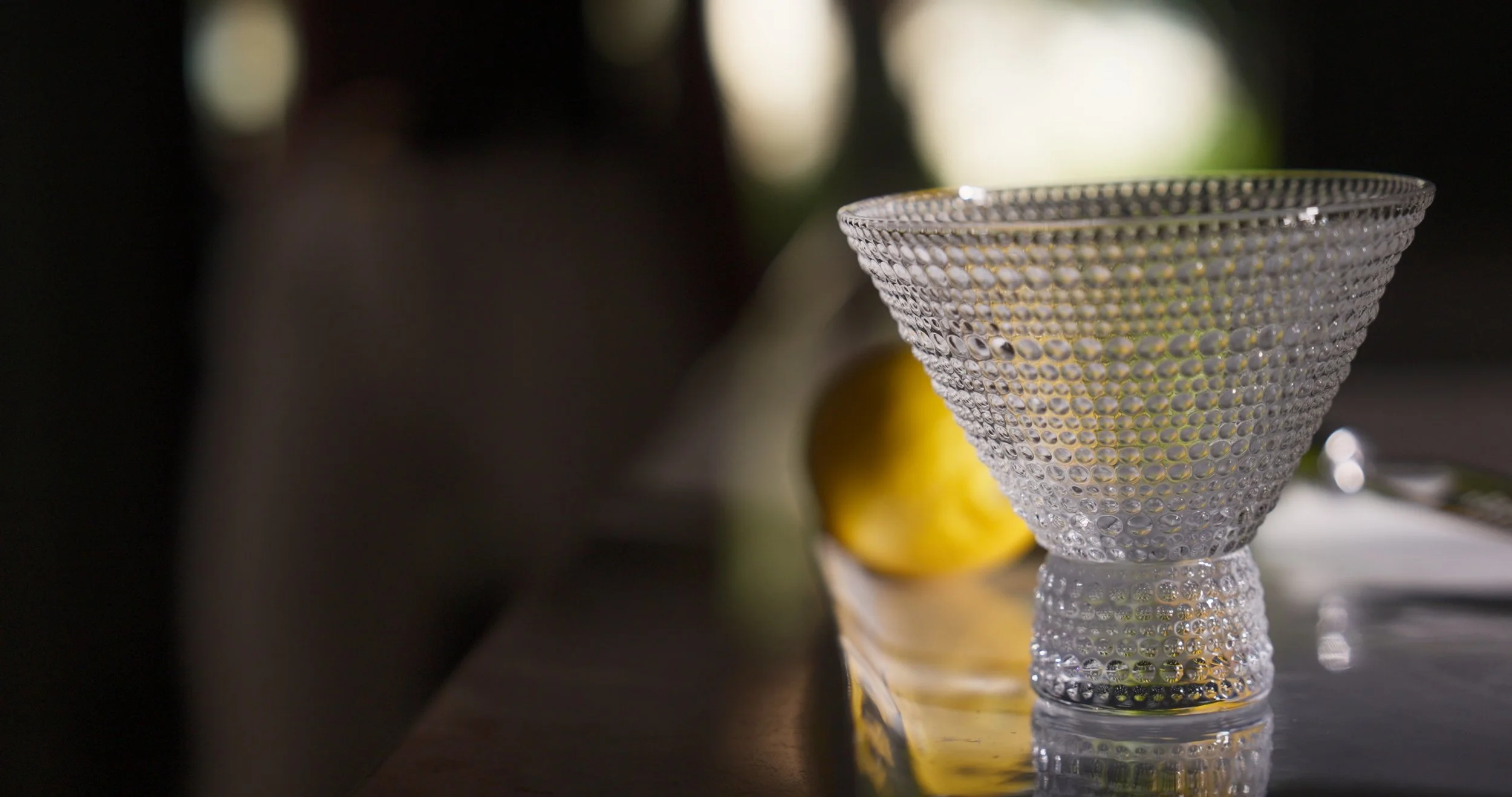 A textured glass bowl with a beaded pattern sits on a reflective surface, with a blurred lemon and utensils in the background.