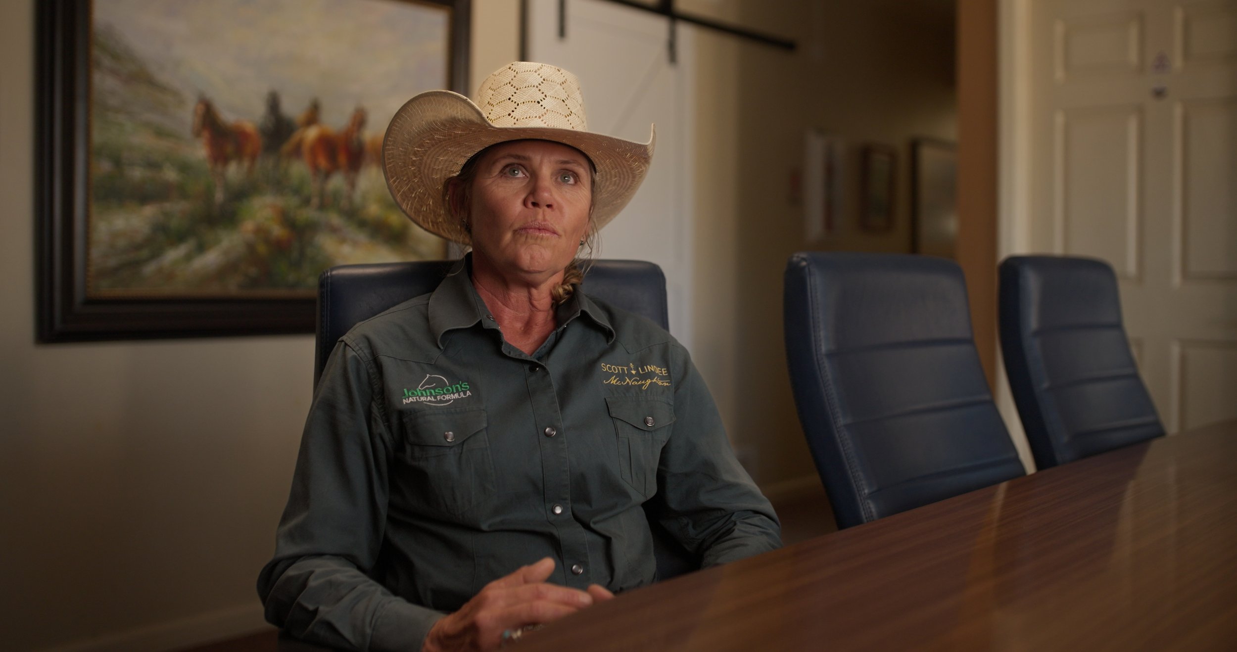 A woman wearing a straw cowboy hat and a gray button-up shirt with logos, sitting at a wooden conference table in a room with blue chairs and paintings on the wall.