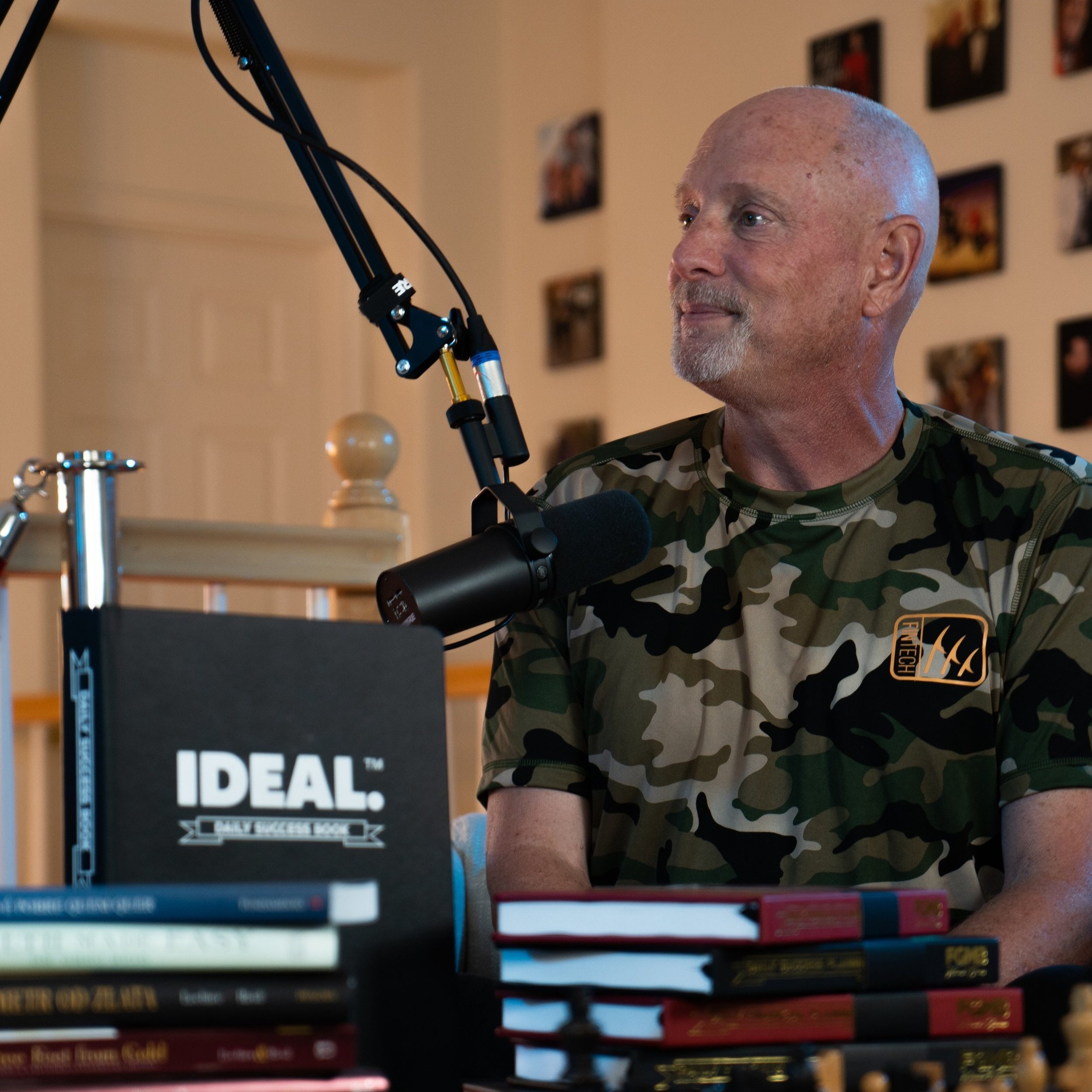 A man with a bald head and a gray beard is sitting at a table in front of a stack of books. He is wearing a camouflage-patterned shirt and is focused on something in front of him. Part of a black microphone setup is visible in the foreground, and a w