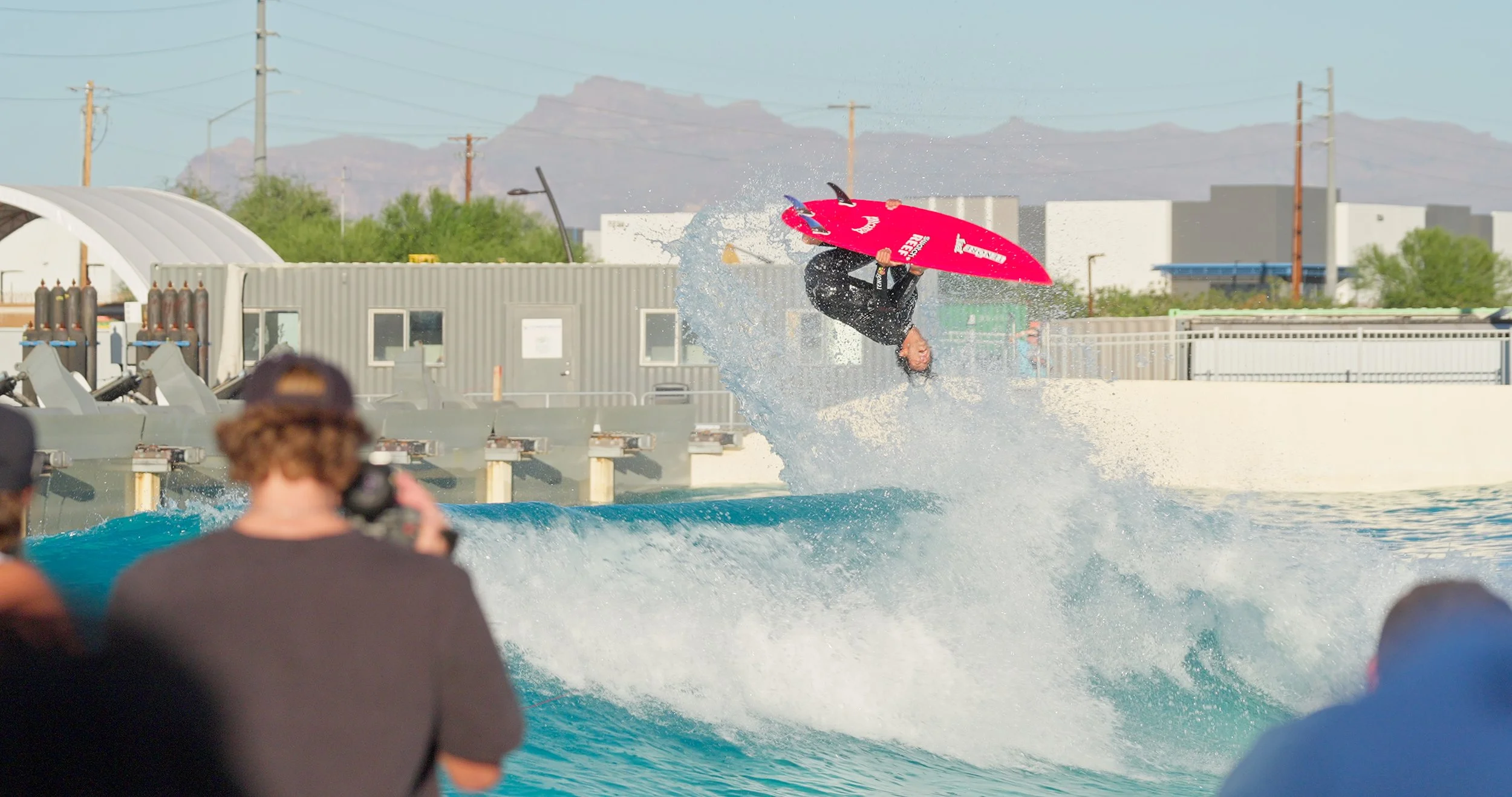 A person performing an aerial flip on a pink surfboard in an artificial wave pool, with onlookers watching.