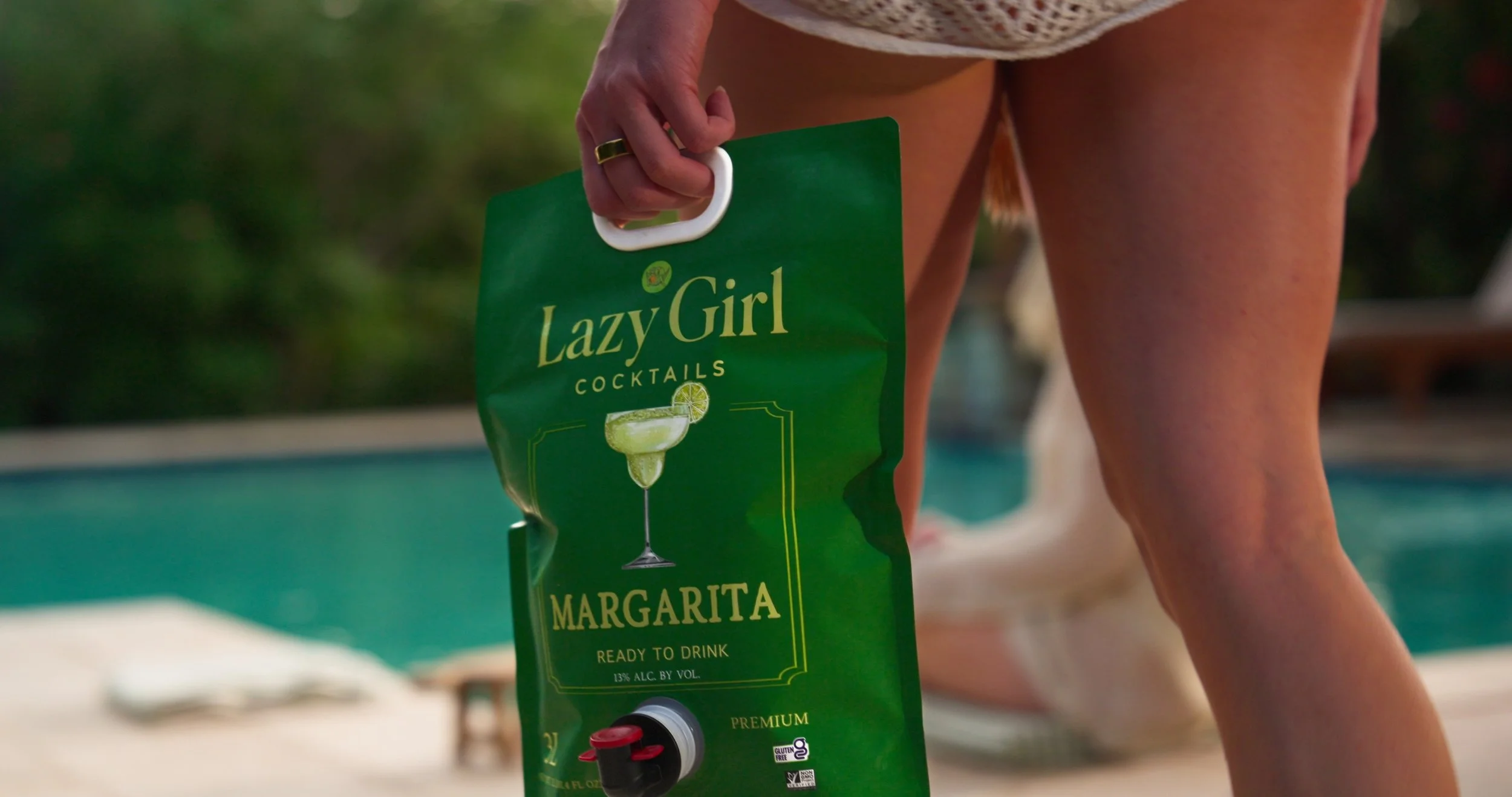 Person holding a green Lazy Girl Margaritas cocktail pouch beside a swimming pool, with part of their leg visible, and a blurred poolside background.