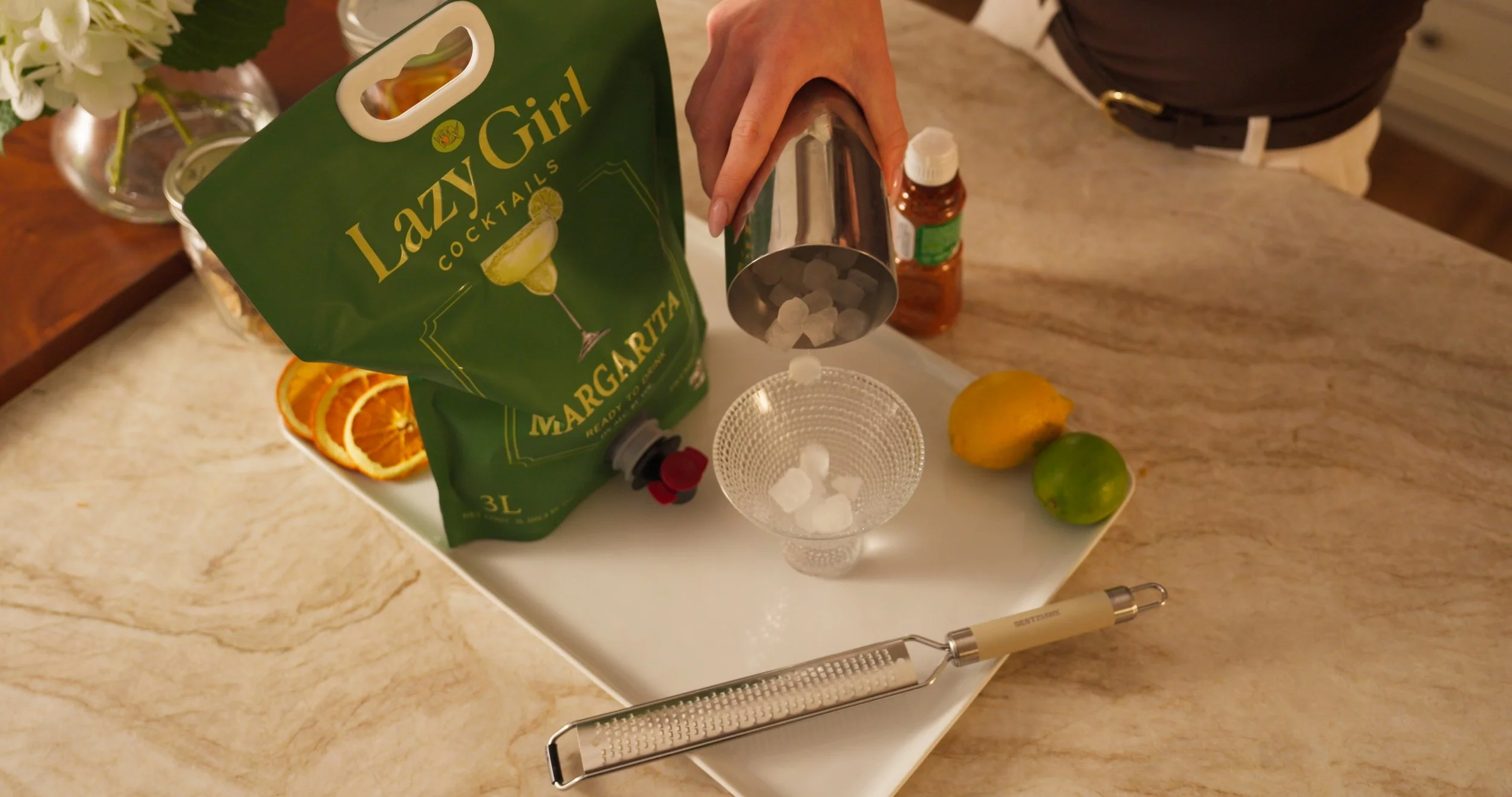 A person is pouring large ice cubes into a glass, with a bag of Lazy Girl Margaritas mix, a lemon, a lime, and a bottle of alcohol nearby on a marble countertop.
