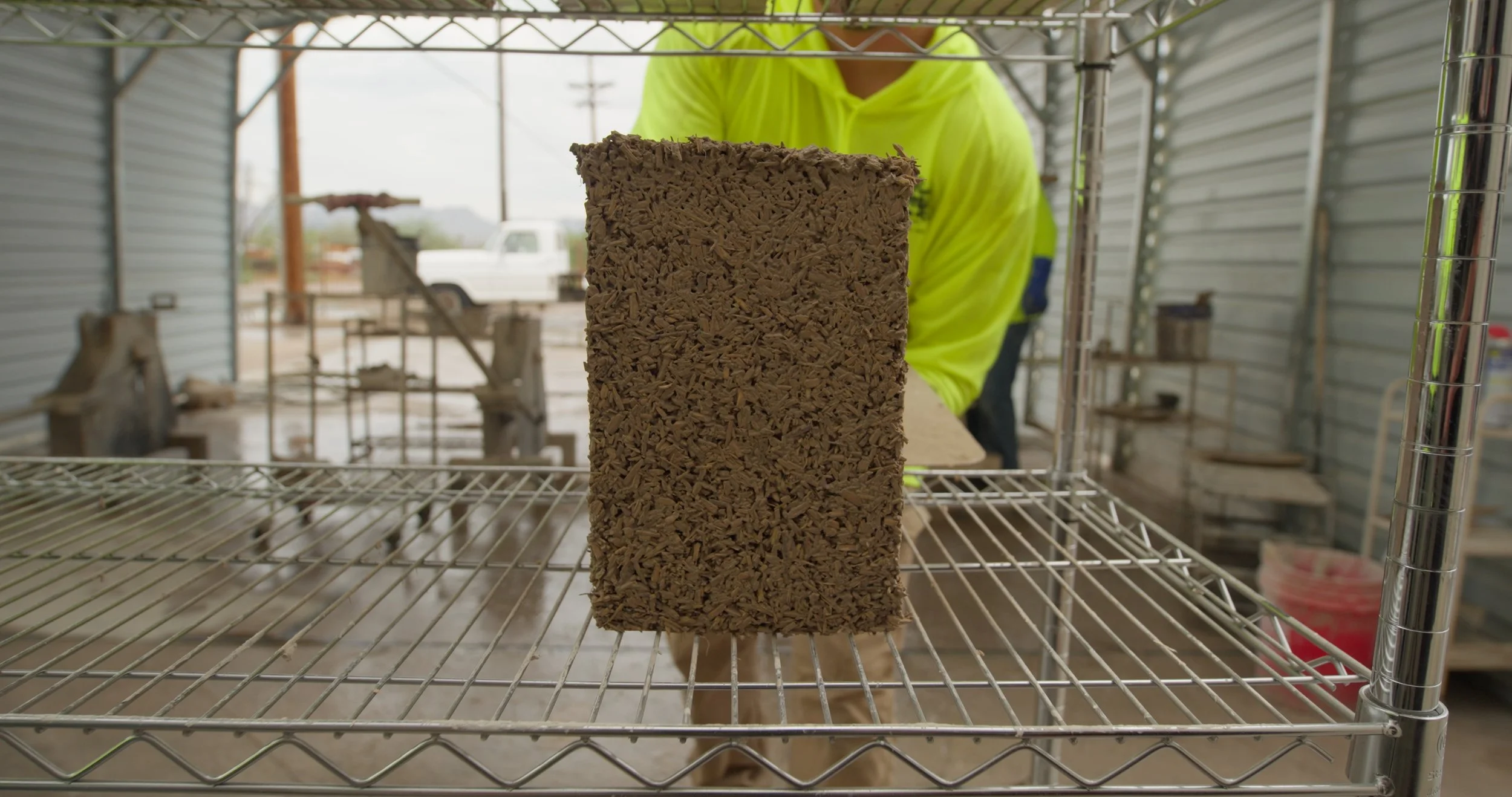 A person in a bright yellow shirt working on a wood project inside a metal workshop, viewed through a metal shelving unit with a rectangular block of wood or particle board in the center.