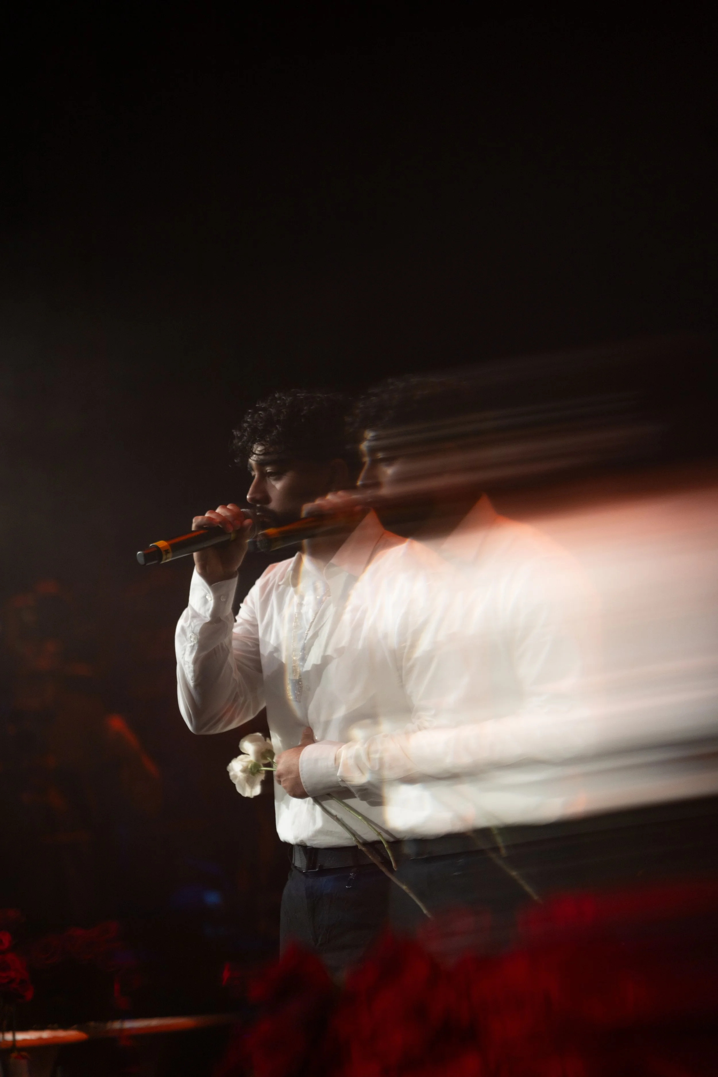 A man holding a microphone with a motion blur effect, wearing a white shirt, standing in a dark setting, possibly a stage, holding a white flower.