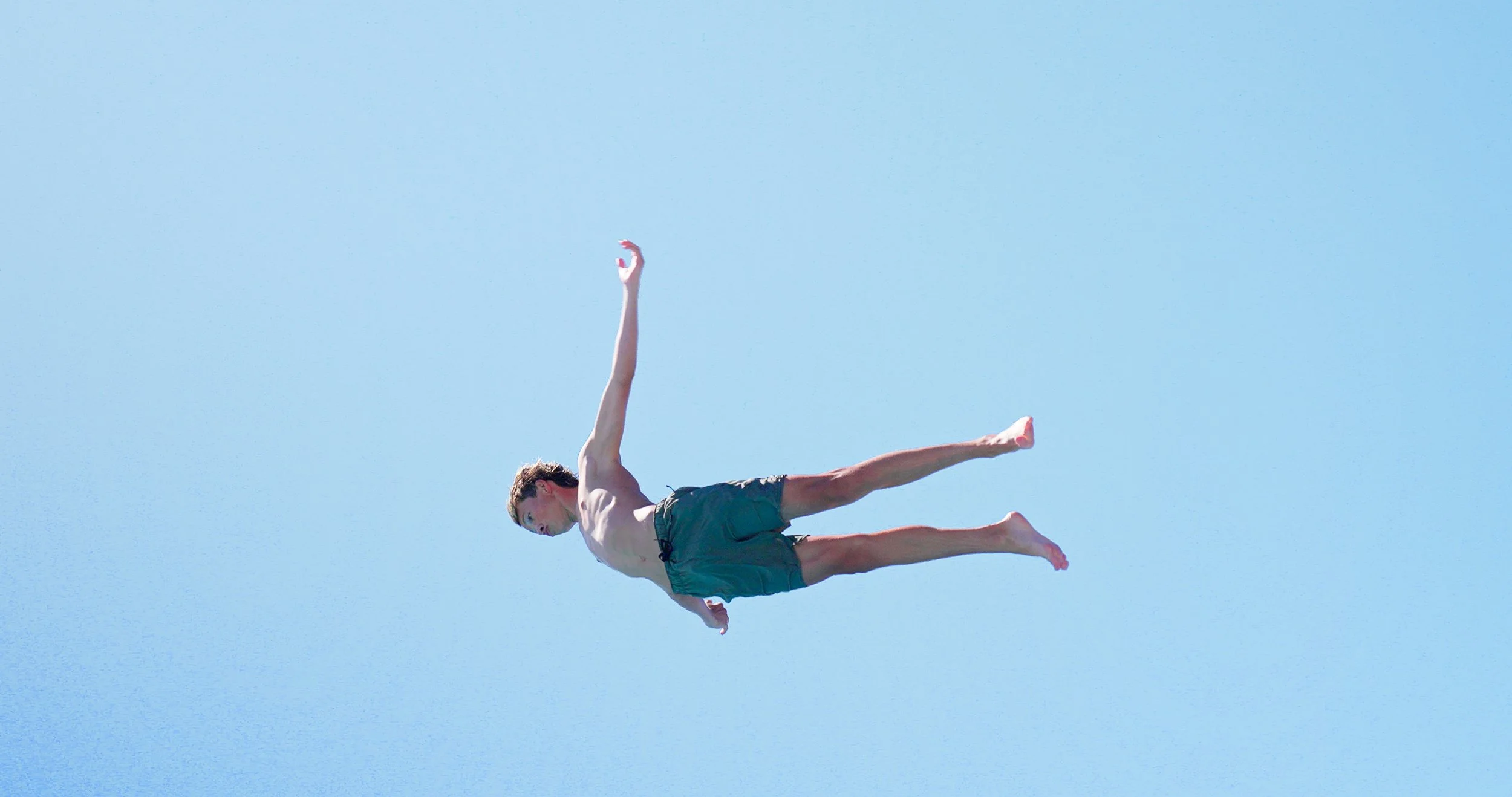 A young man in swimming trunks is diving into the water, with his body fully extended and arms reaching upward against a clear blue sky.