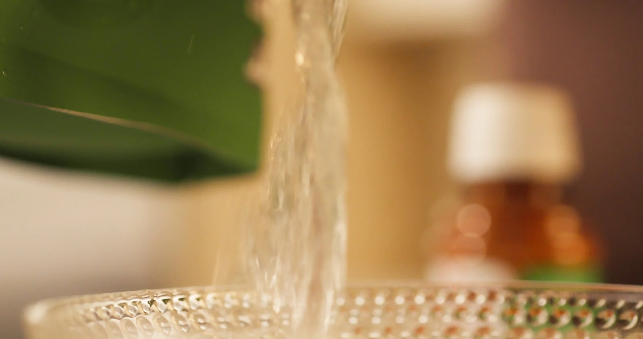Close-up of a glass bowl with a decorative pattern, with water pouring into it from above, and a blurred background with a lamp and a green leaf to the left.