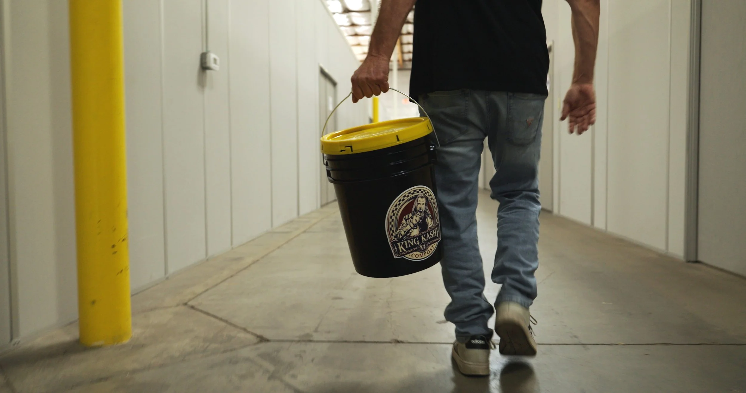A person walking down a hallway carrying a black bucket with a yellow lid, labeled 'King Kashi Compost'.