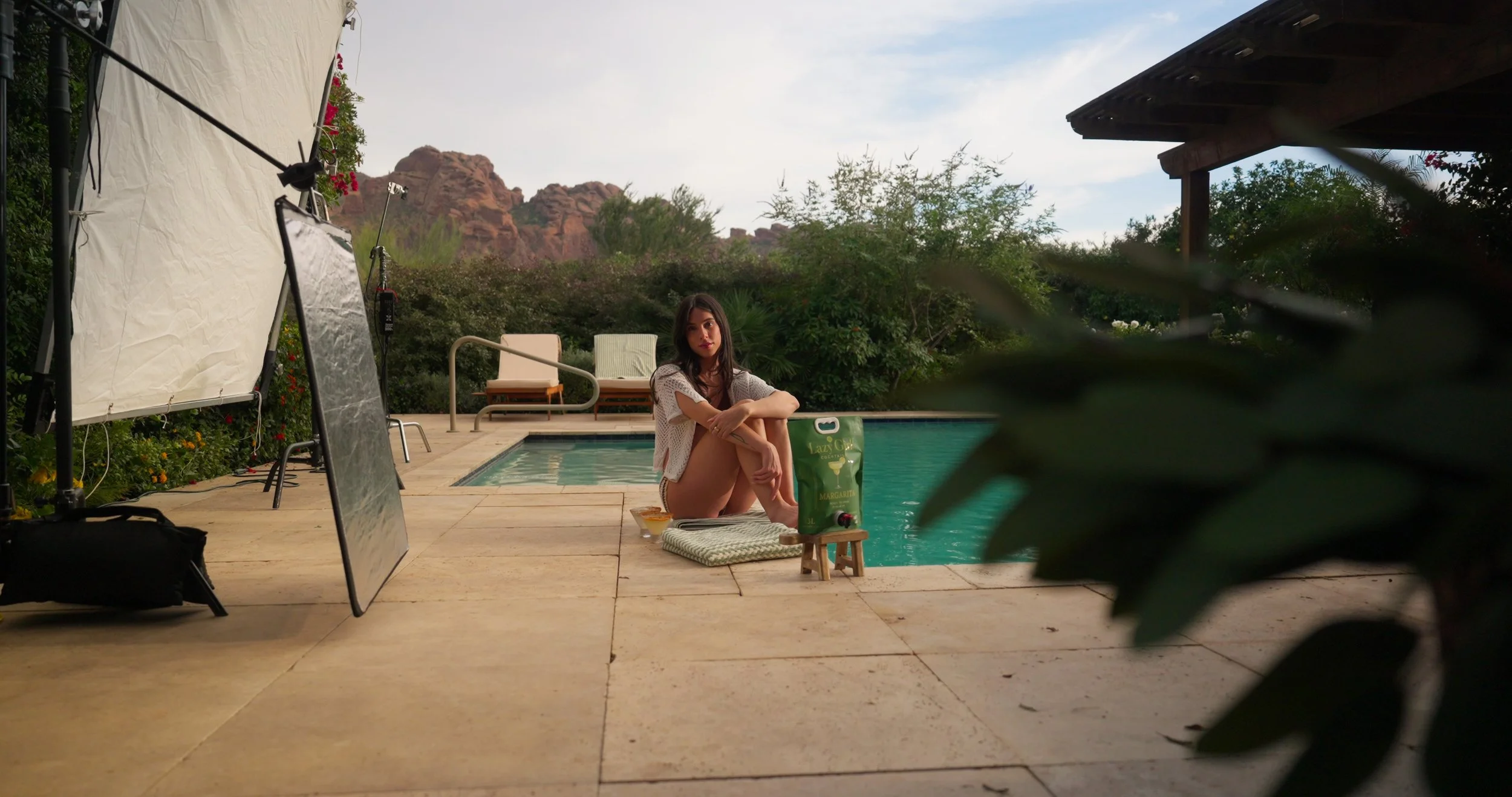 A young woman sits on the poolside with her knees up, surrounded by a small lounge area, greenery, and a mountain backdrop. There is professional photography and lighting equipment nearby, indicating a photo shoot is in progress.
