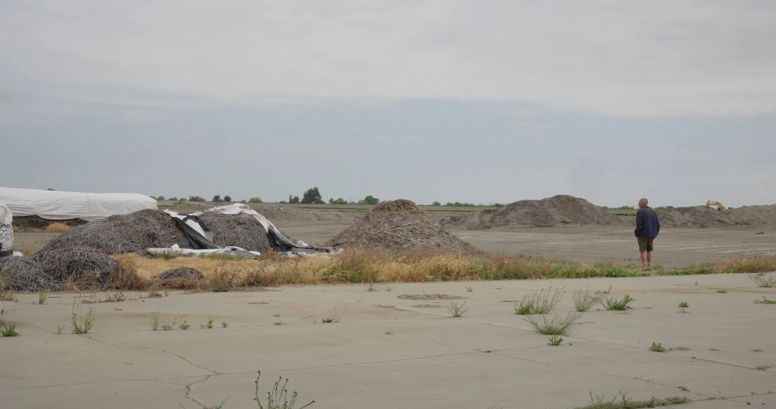 A person stands on cracked pavement looking at mounds of dirt and rocks in a barren, open landscape under a cloudy sky.