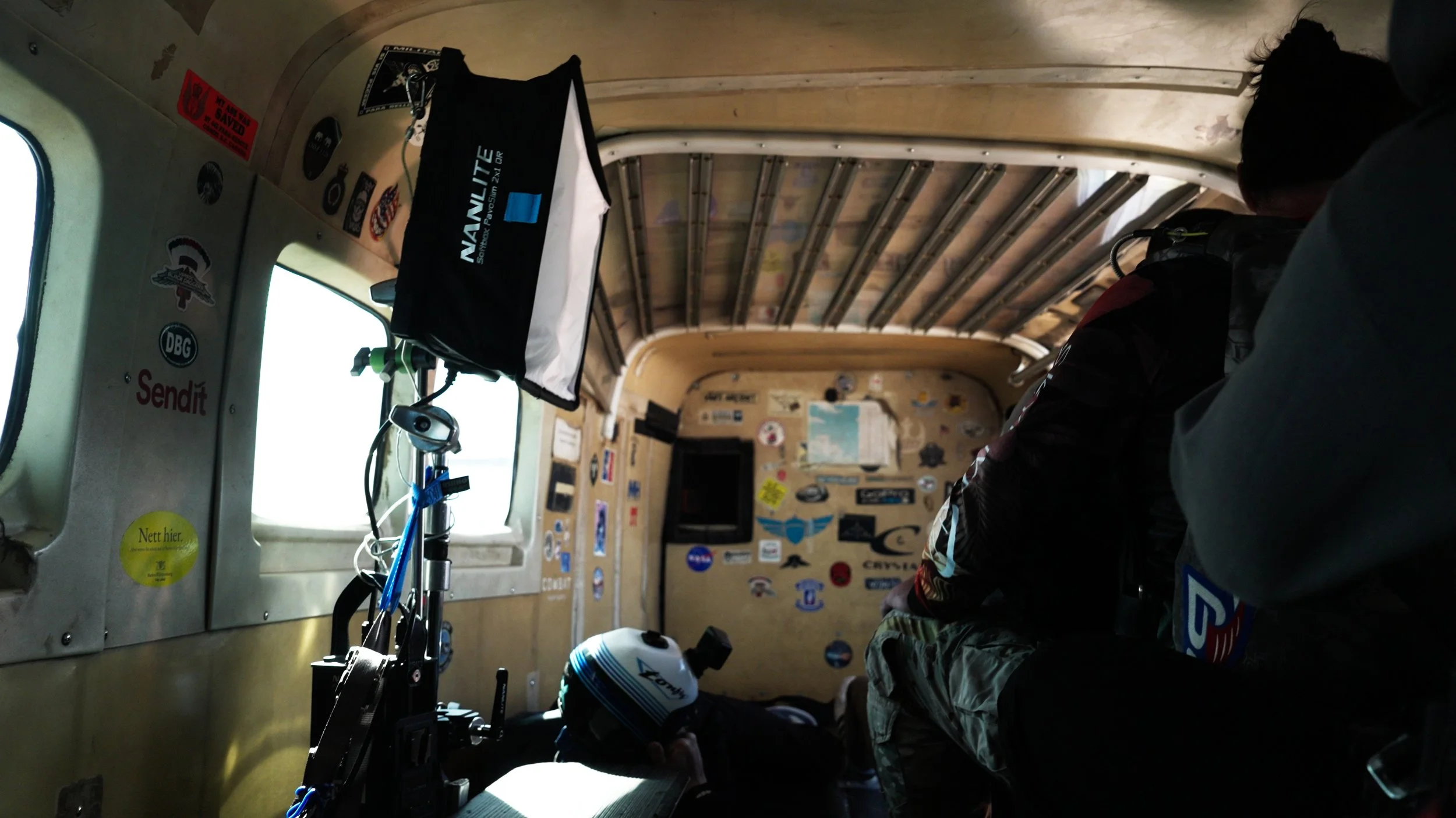 Interior of a small aircraft with seating on the right side, walls adorned with numerous stickers and decals, a small abyss mounted on the left, and windows showing a bright sky outside.