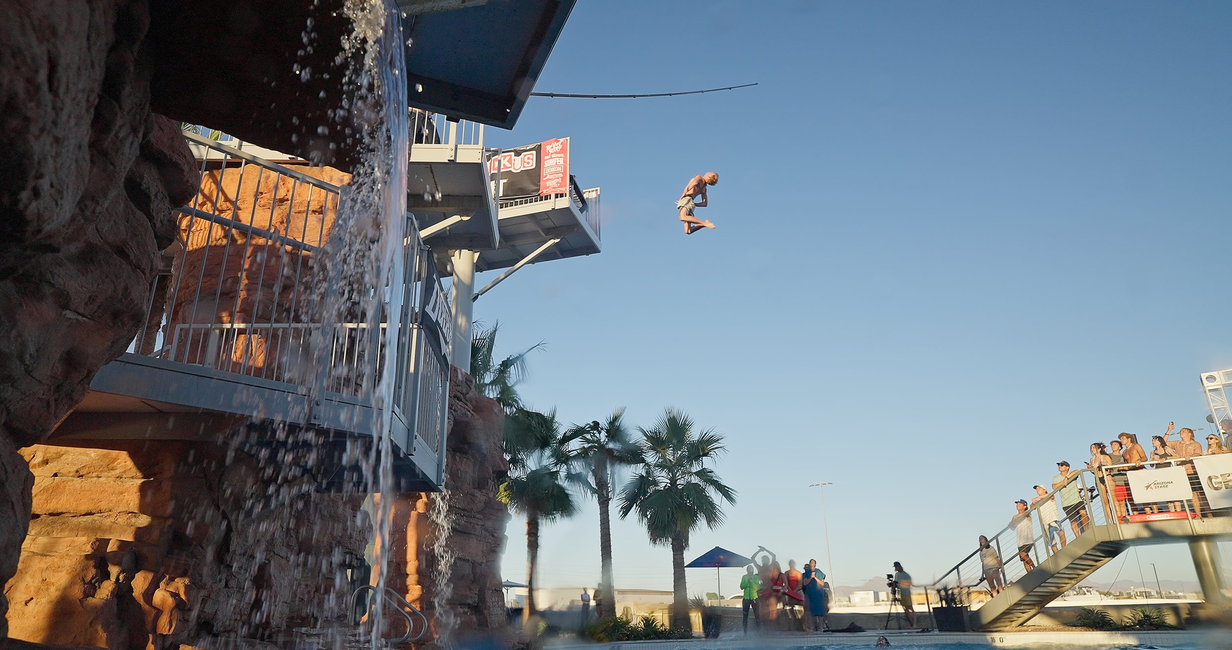 A person mid-air after jumping from a diving board at a water park, with spectators watching from a platform, palm trees, and a clear blue sky in the background.