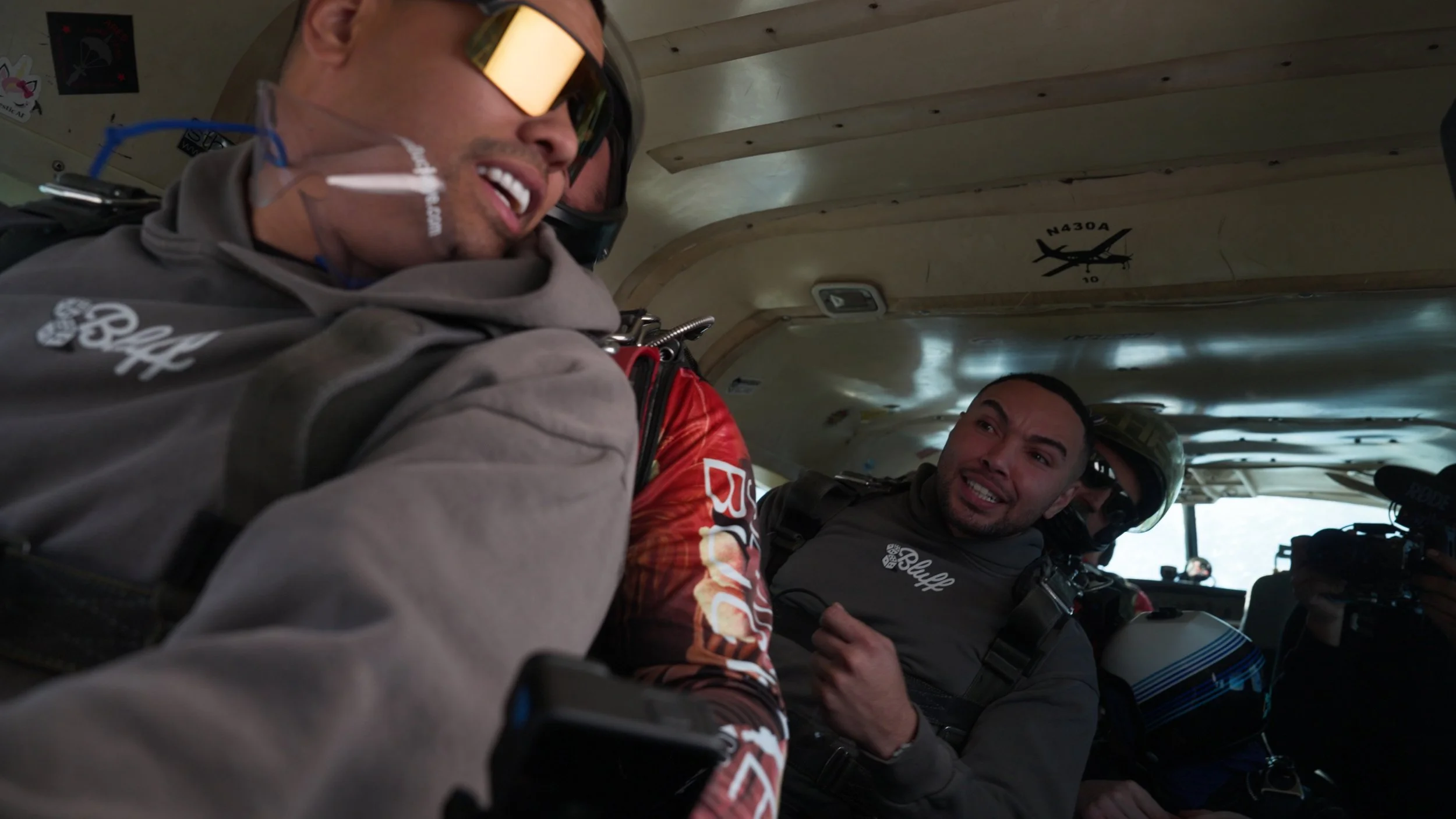 Three skydivers inside an aircraft with a curved ceiling, preparing for a jump; one wearing gold-tinted goggles and a gray jumpsuit, another with a red and black jumpsuit and a helmet, and the third with a gray jumpsuit and a black helmet.
