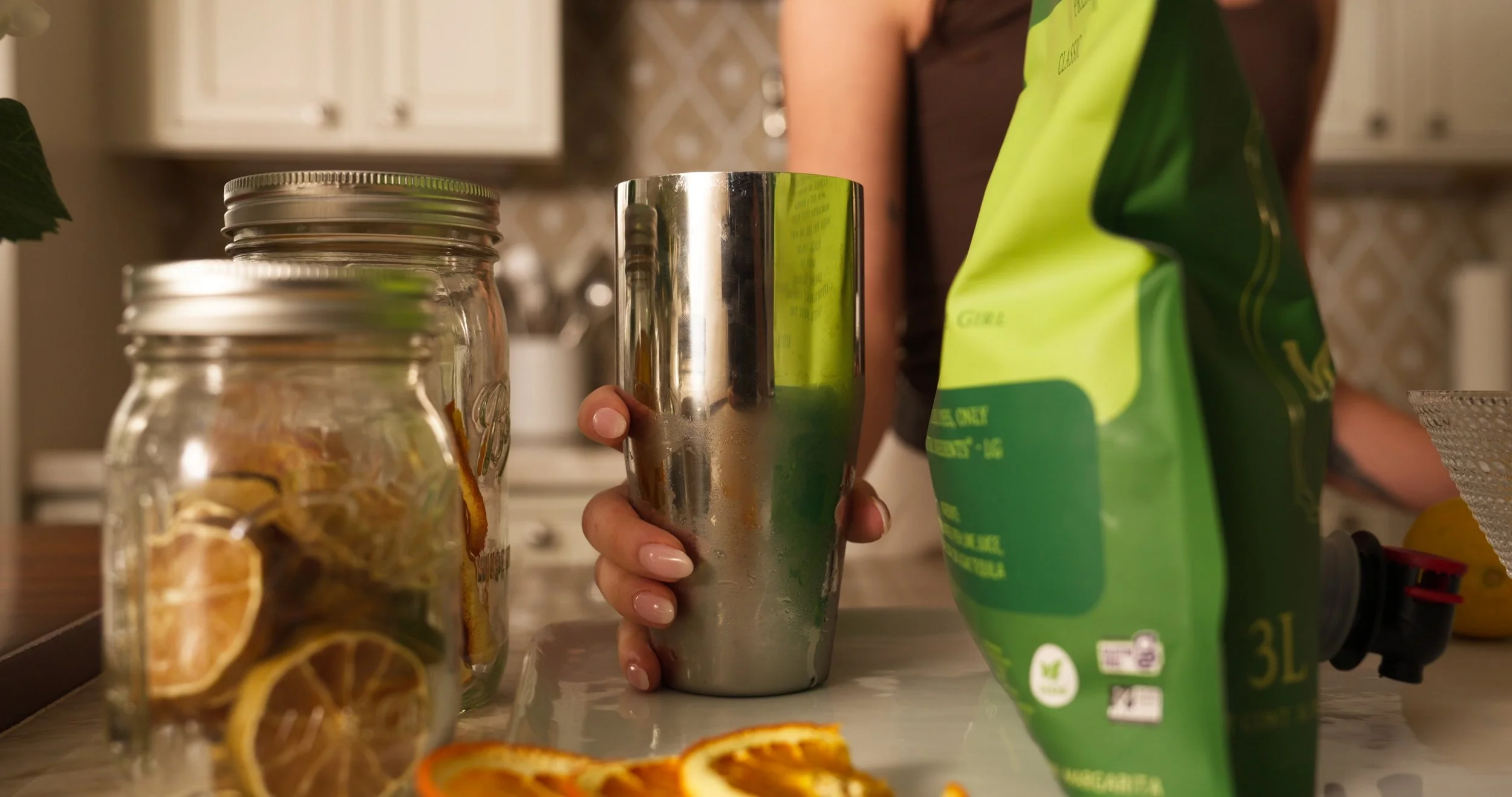 A person is holding a frosted metal tumbler glass on a kitchen counter, with jars of dried citrus slices and orange peel nearby. A green bag of dried citrus slices is also on the counter.