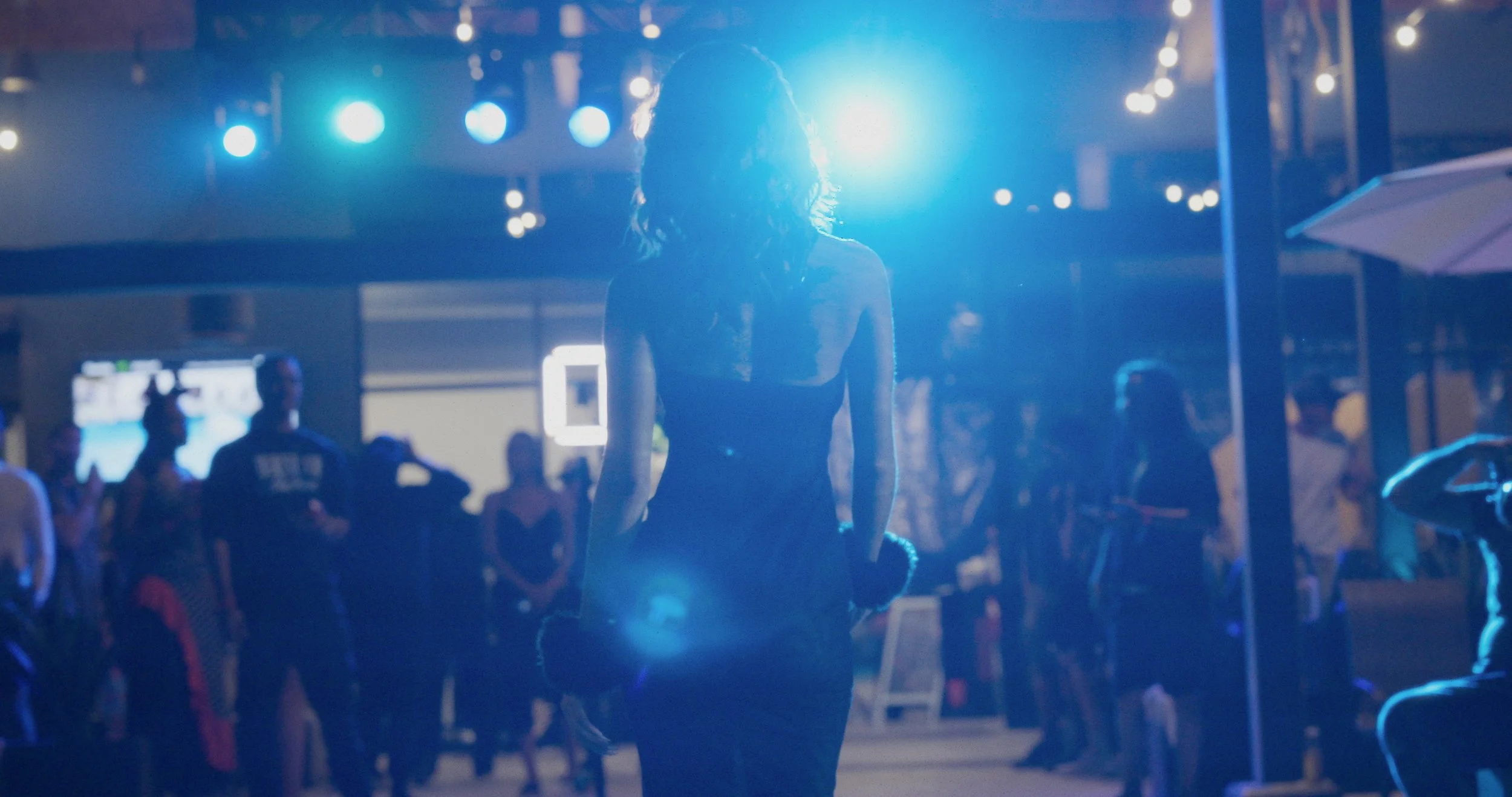 Backlit woman with curly hair at a night event, surrounded by people and illuminated by blue stage lights.
