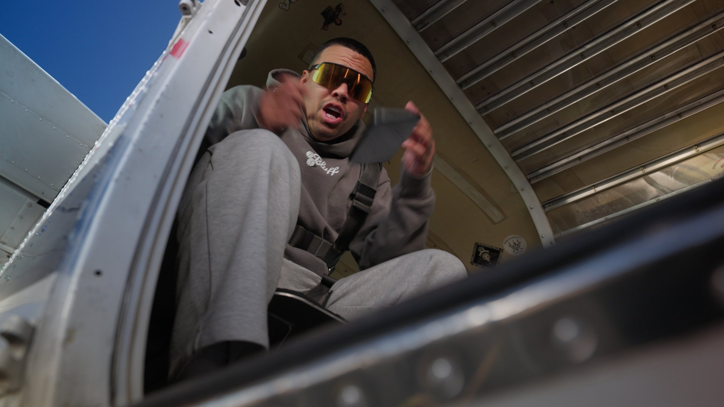 A man sitting inside an aircraft, wearing sunglasses and holding a tablet, looking up towards the camera with a surprised expression.