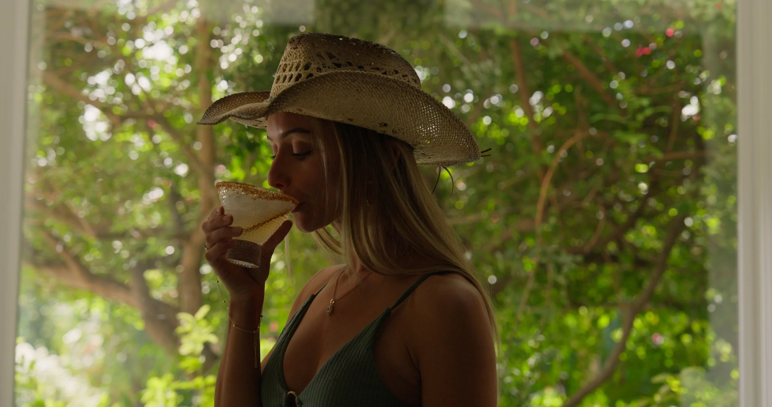A woman wearing a wide-brimmed straw hat and a dark sleeveless top, holding a margarita glass near her lips, standing near a window with a background of lush green trees.