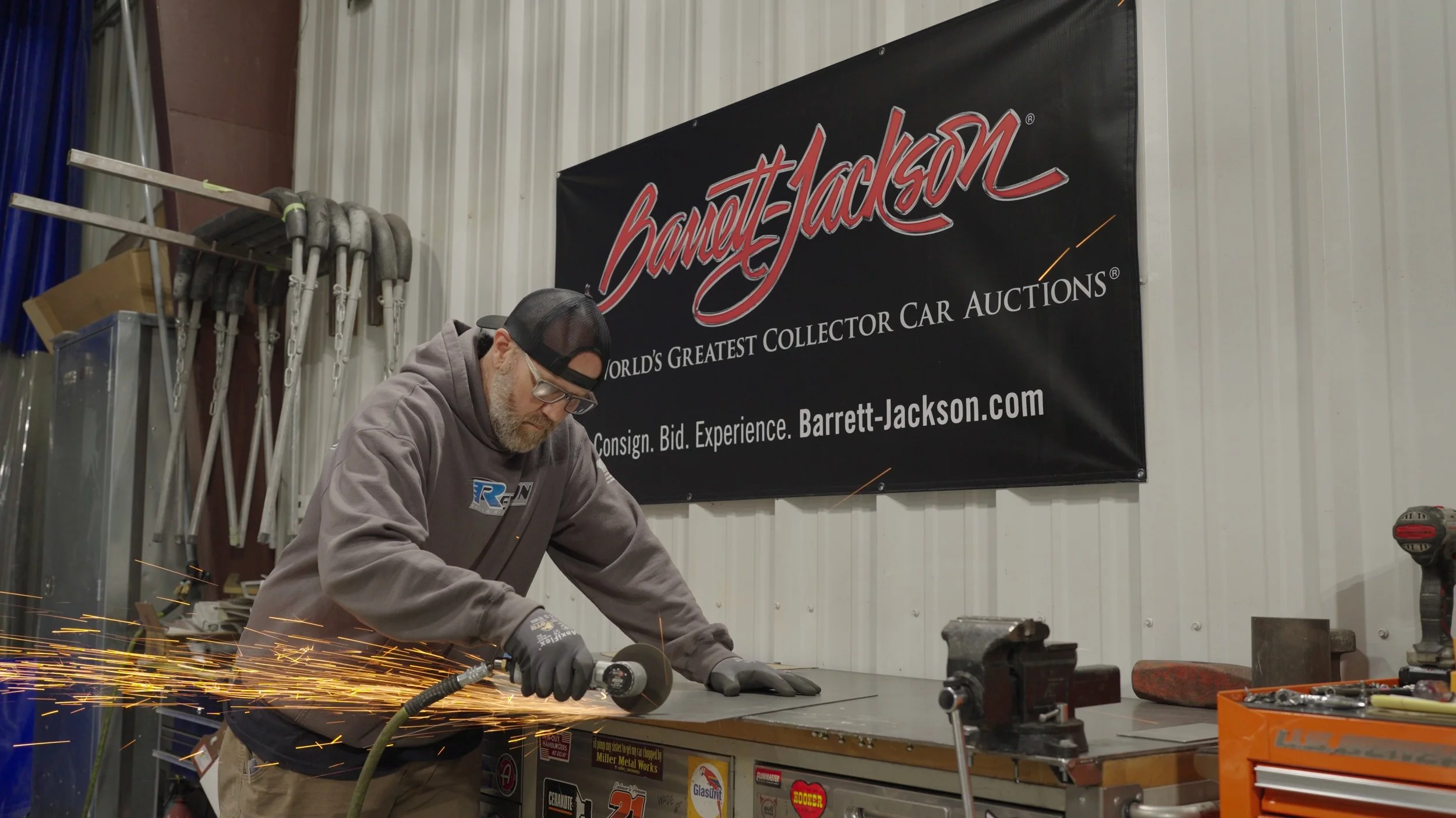 Custom car builder grinding steel in workshop with Barrett-Jackson banner visible during documentary production.