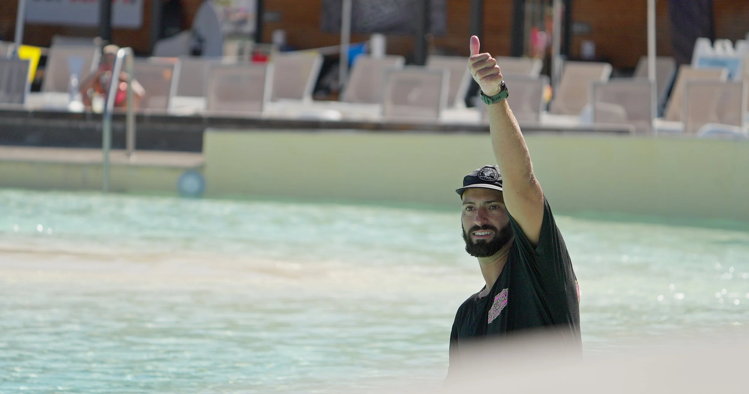 A man with a beard and black t-shirt giving a thumbs up while standing in the water at a beach or pool.