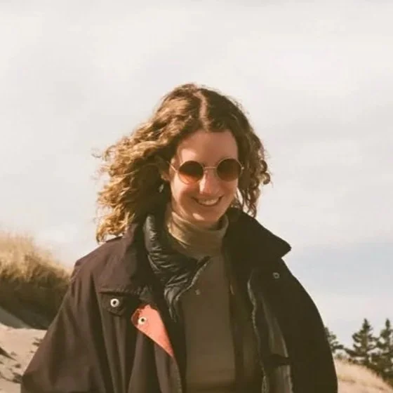 A young woman with curly hair smiling outdoors in natural light, wearing a black shirt, red patterned dress, and cream-colored cardigan.