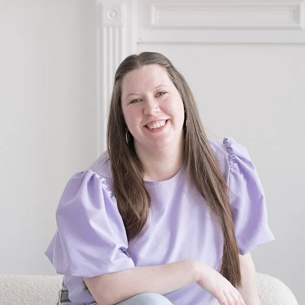 A woman with light skin and brown hair tied with a pink ribbon, smiling, wearing a white knitted sweater, standing in front of a plain light-colored wall.