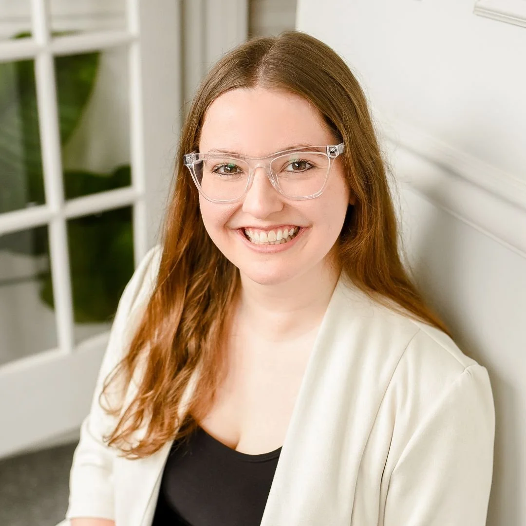 A young woman with long, wavy red hair wearing clear glasses and a light-colored blazer, smiling while sitting indoors.