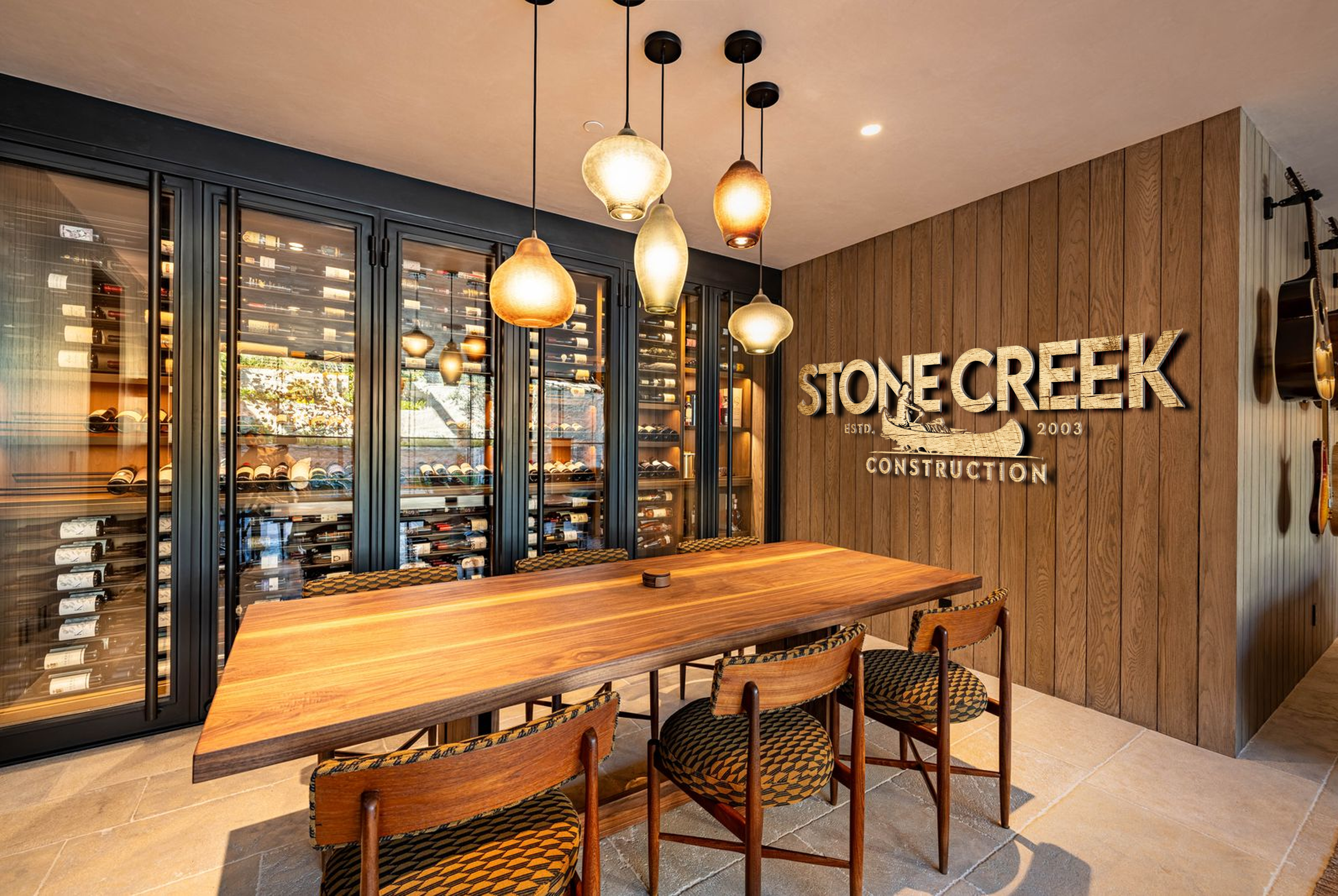 Interior of a wine tasting room with a wooden table, patterned chairs, a wine bottle display in a glass cabinet, and hanging pendant lights, with a Stone Creek Construction sign on the wooden wall.