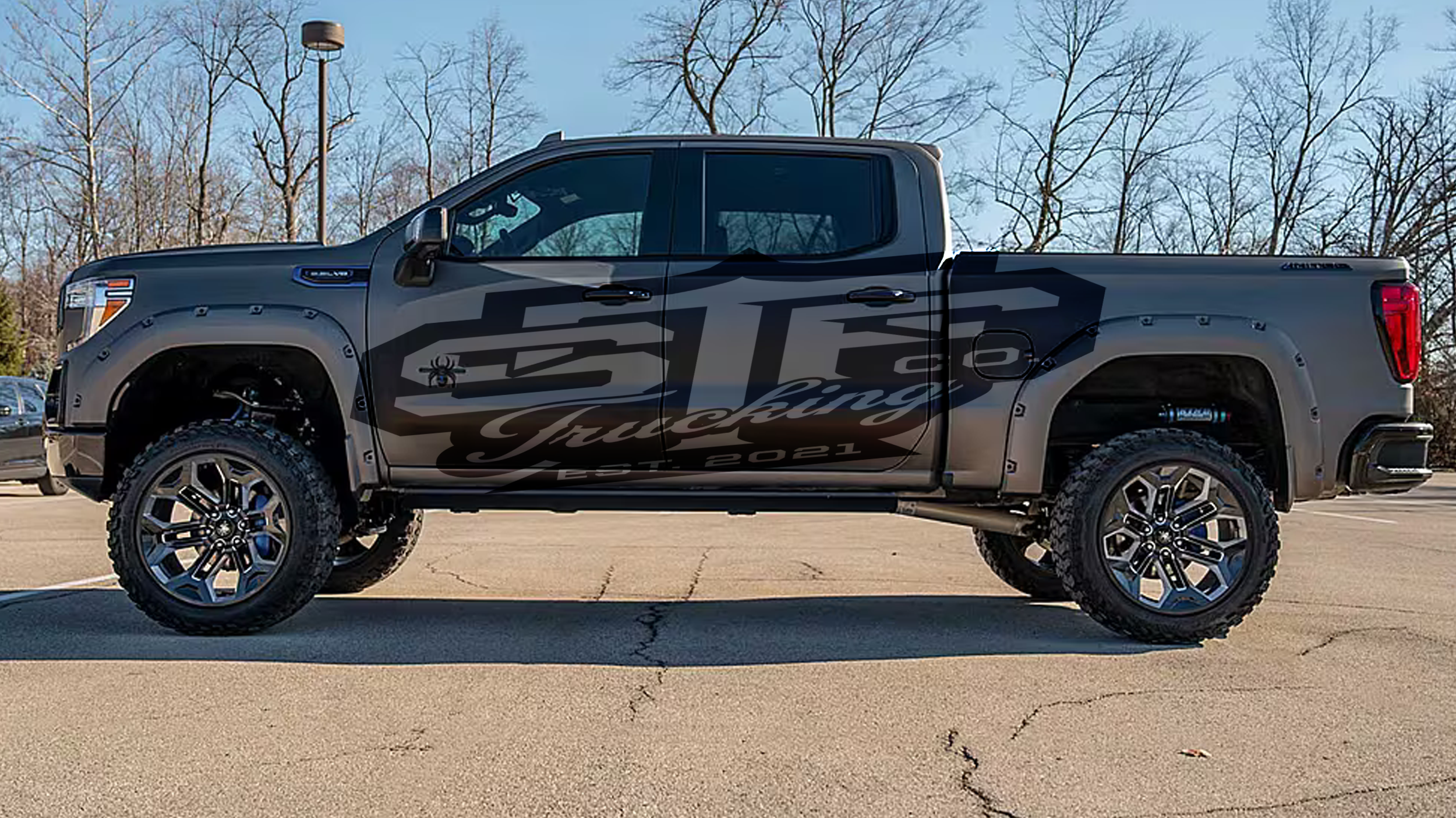 A vehicle wrap of black pickup truck with large tires parked in a lot, featuring custom graphics and stickers on the body, under a clear blue sky with leafless trees in the background.
