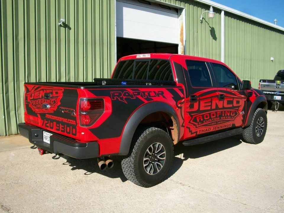 Red and black truck with Jenco Roofing logo and branding parked outside a green building with a roll-up door.