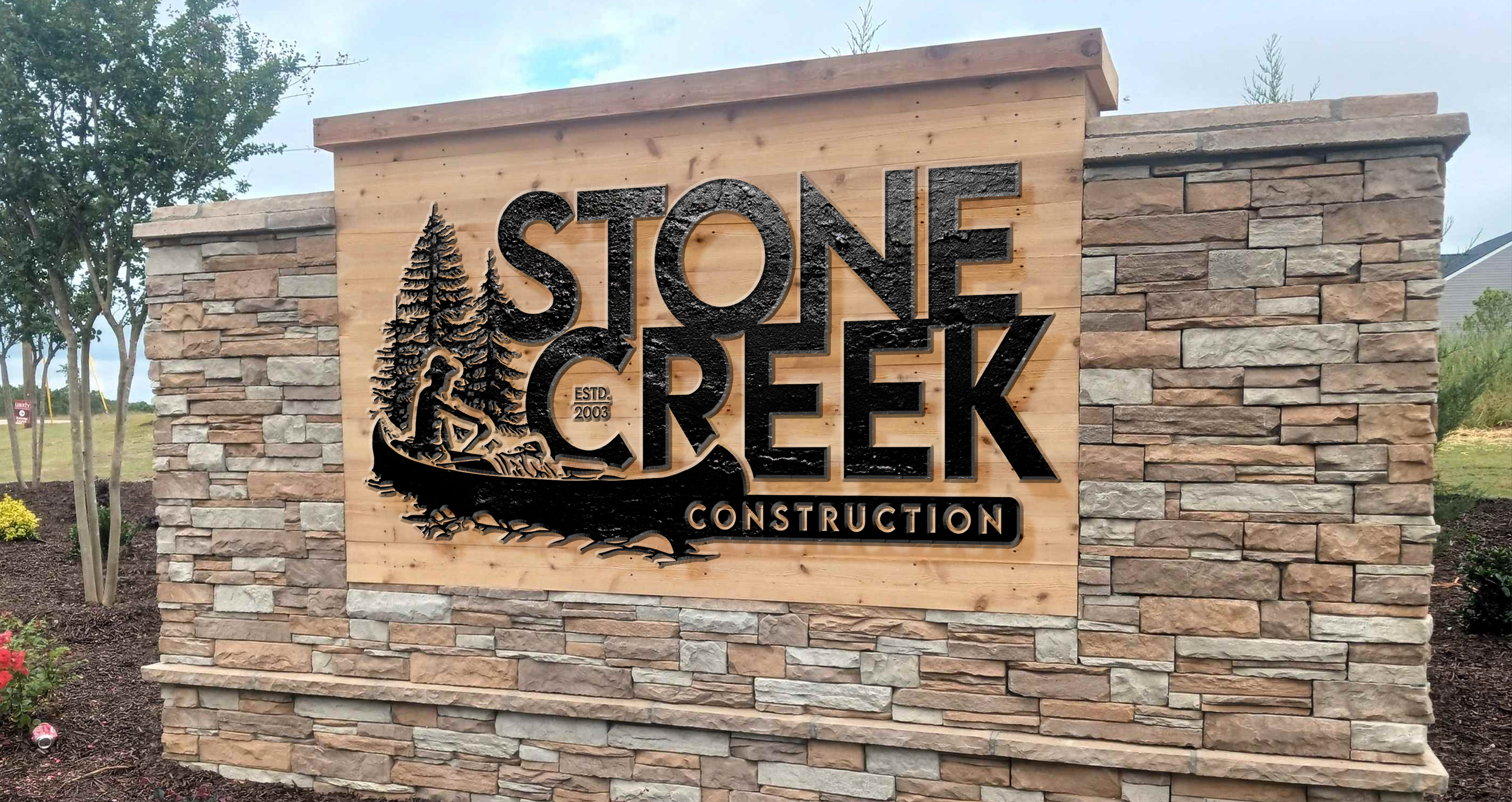 A stone and wood sign reads 'Stone Creek Construction,' with an illustration of a woman sitting by trees, established in 2003, set against a landscaped area with plants and trees.