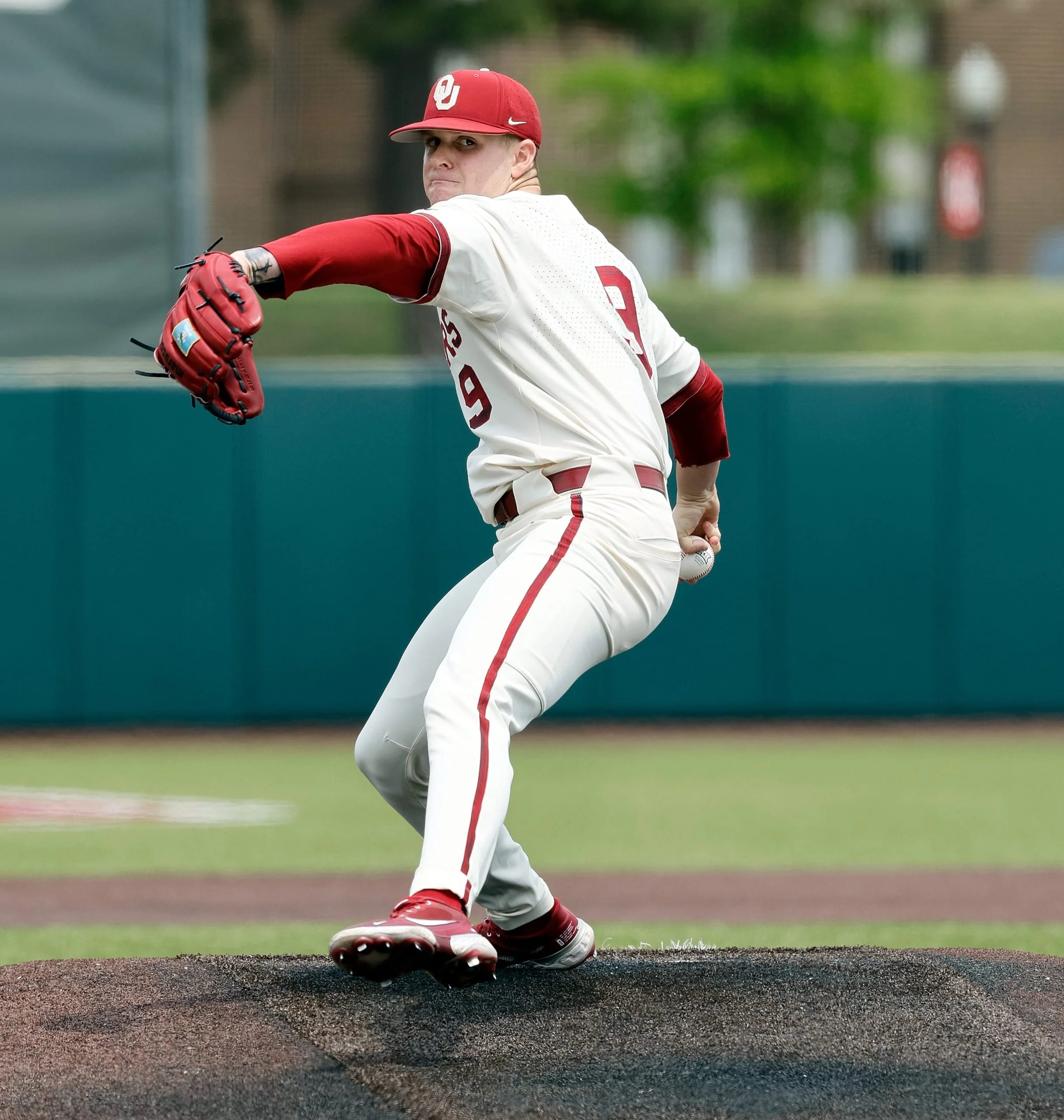 A baseball player dressed in a white and red uniform is pitching on the field, with a green outfield and trees in the background.