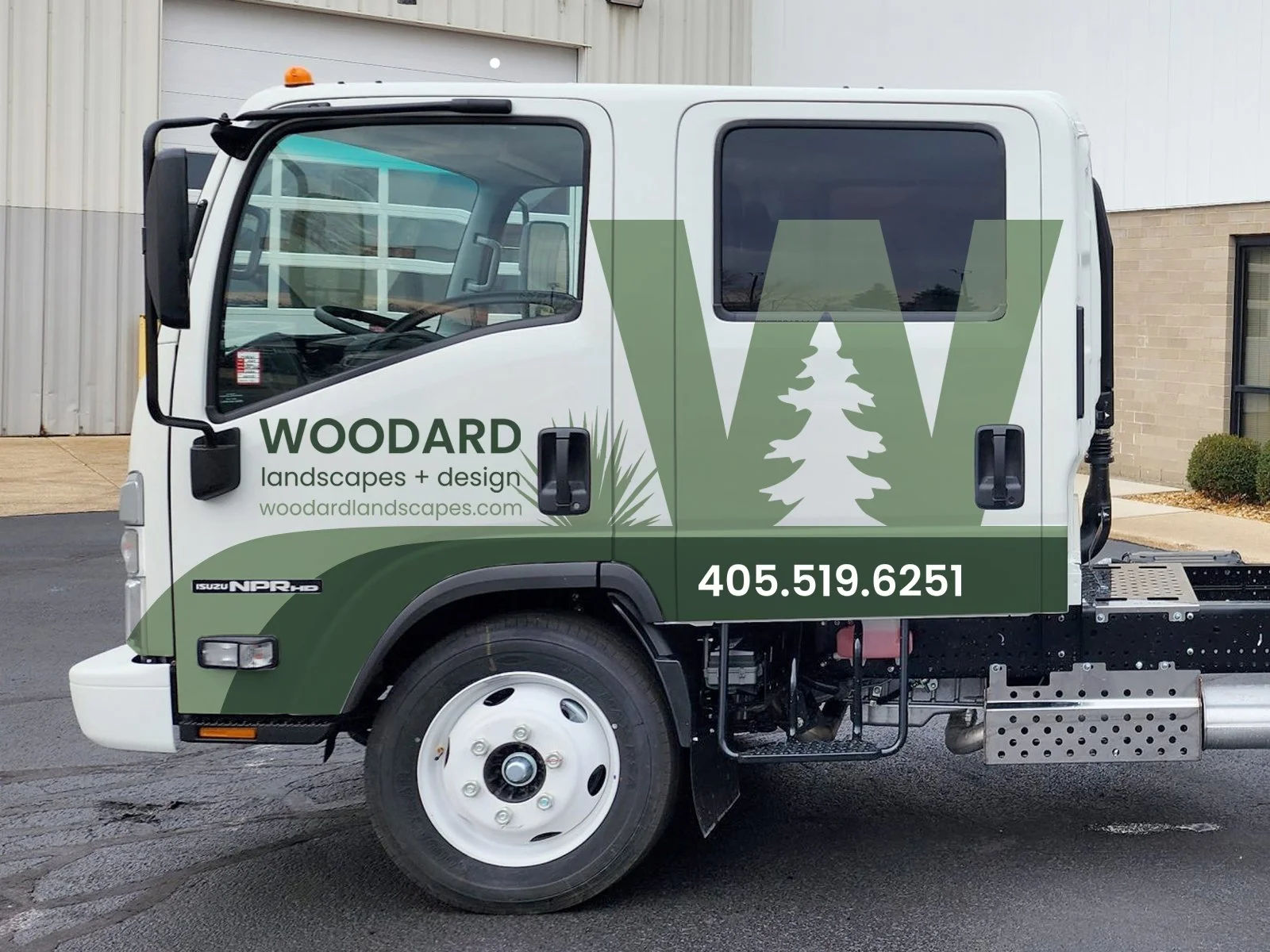 A white truck with green and black vinyl signage for Woodard Landscapes & Design, featuring tree graphics, contact number 405.519.6251, and website woodardlandscapes.com, parked outside a building.