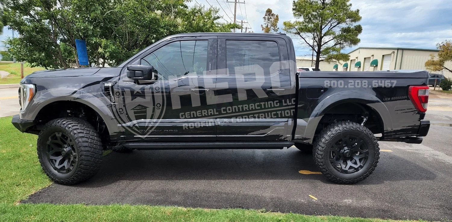 A black pickup truck parked in a parking lot with large off-road tires and a company logo and contact information on the side, advertising general contracting and roofing services. Hero Roofing Company vehicle wrap.