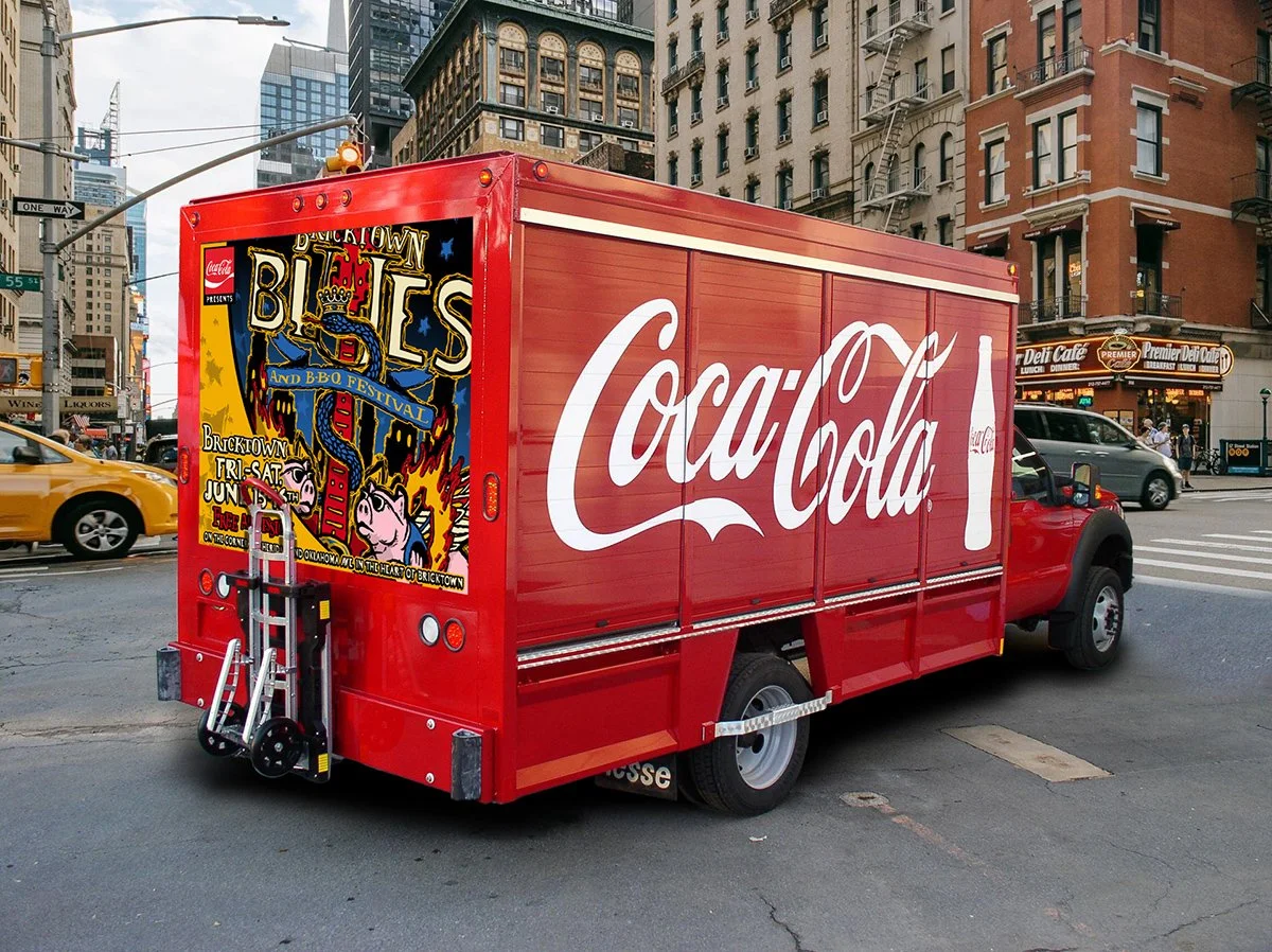 Red Coca-Cola delivery truck parked on city street with skyscrapers and buildings in background. The truck features large white Coca-Cola logo and an advertisement poster for Bricktown Blues and BBQ Festival.