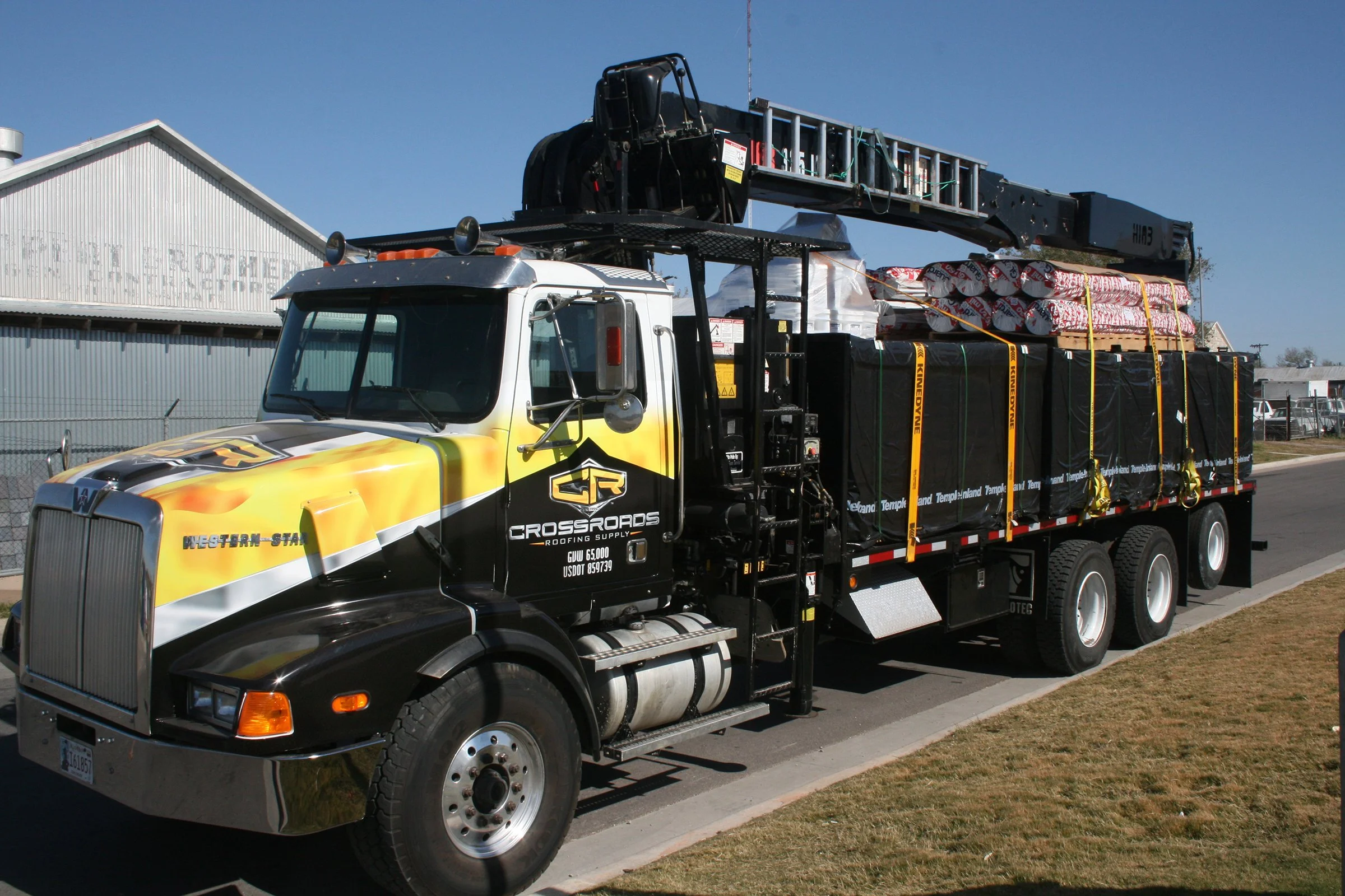 A flatbed truck carrying building materials, including wrapped rolls of insulation and other supplies, parked on the side of a street near industrial buildings under a clear blue sky.