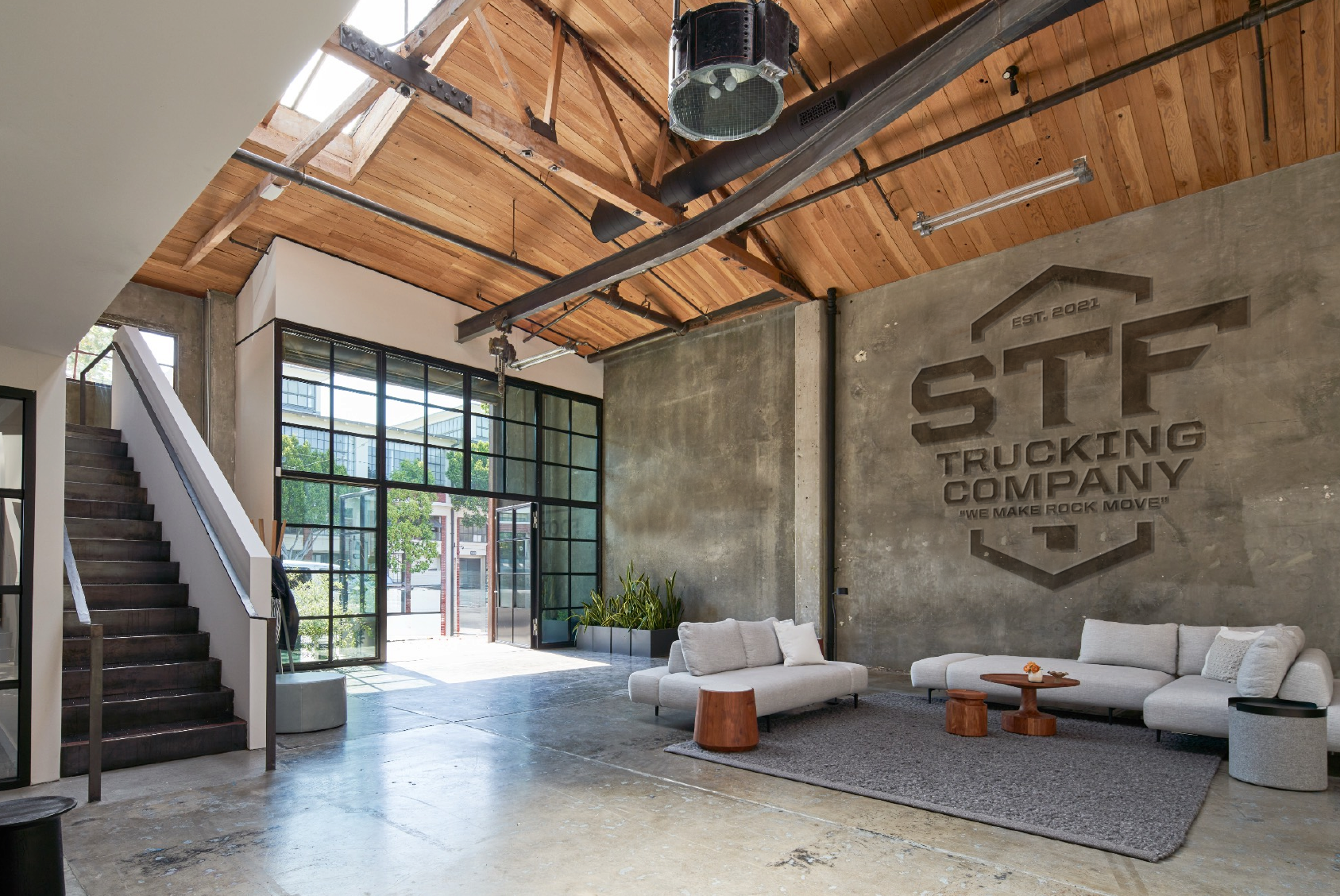 Interior of a modern industrial-style lobby with a concrete wall displaying the logo of ST Trucking Company, wooden ceiling, large window, white sofa, and small round wooden tables.
