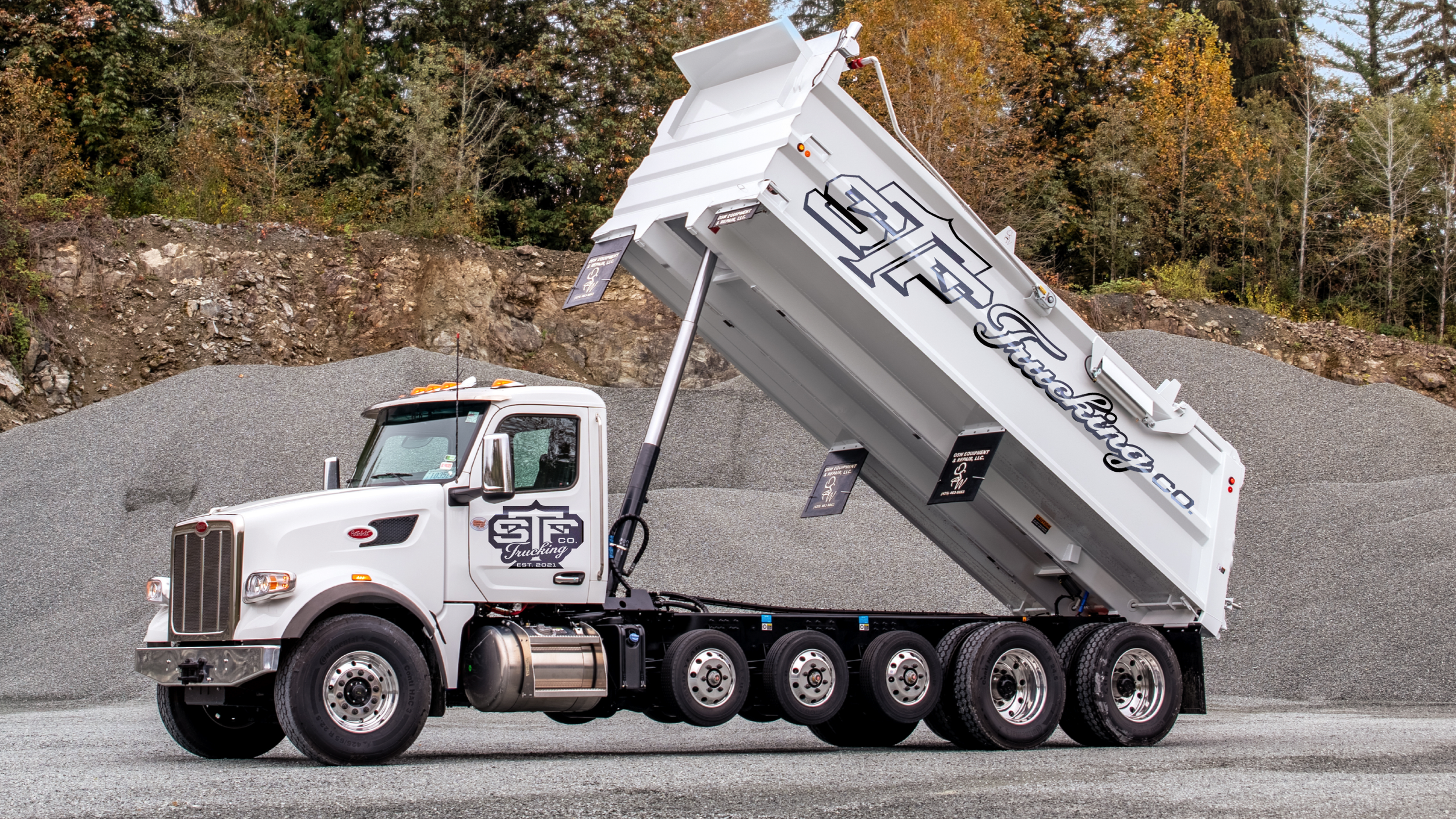A vehicle wrap of a white dump truck with the bed raised, displaying black lettering on the side, parked on a gravel surface with a background of orange and green trees.