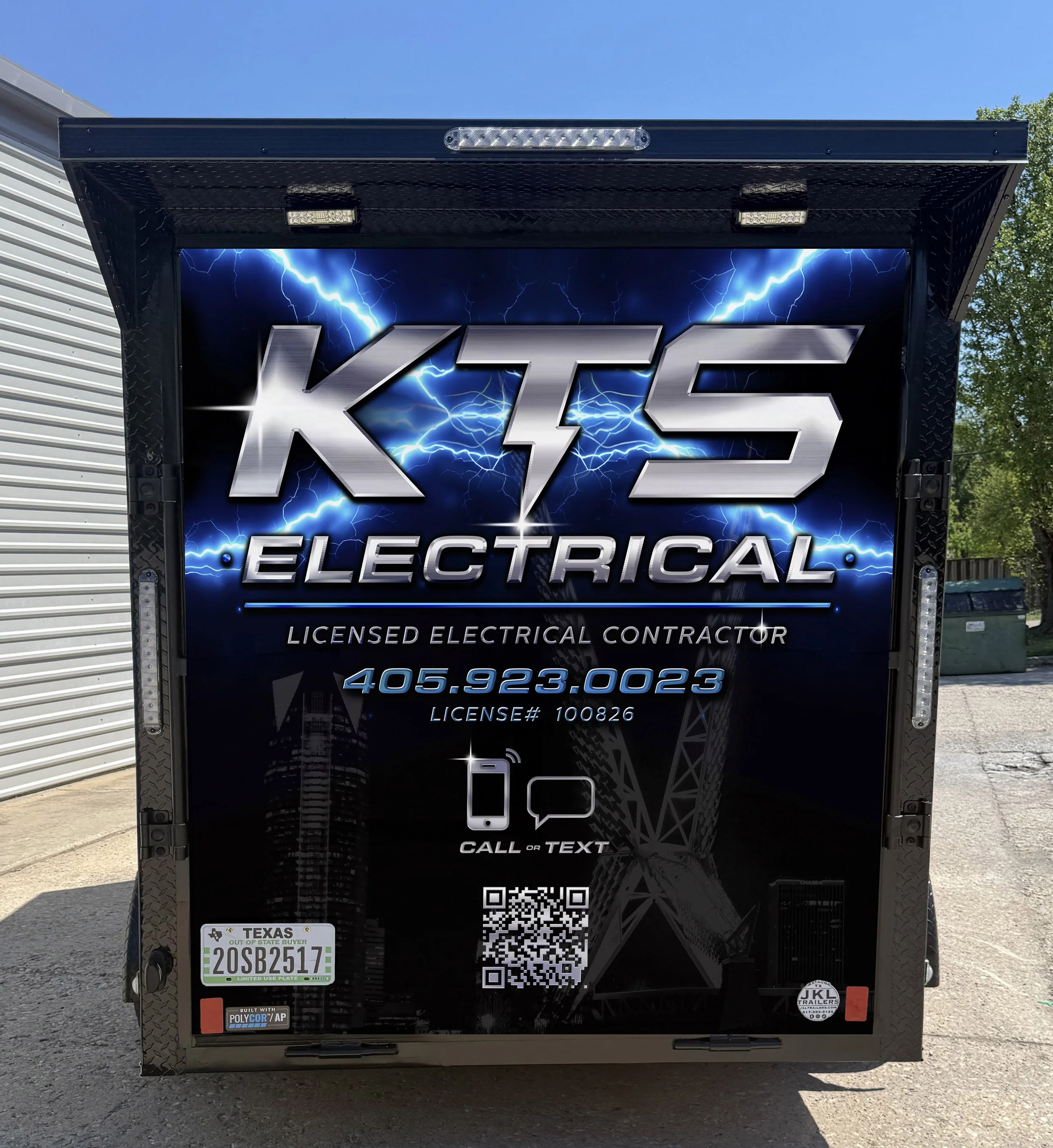 A black trailer with a sign for KTC Electrical, a licensed electrical contractor. The sign features lightning bolts and the company name in metallic letters. Contact information and QR code are visible, along with stickers and logos at the bottom.