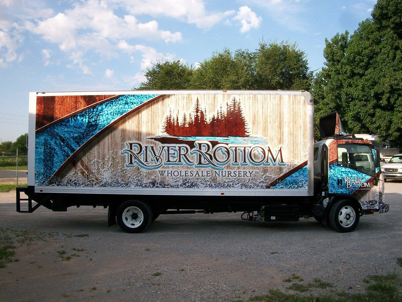 A truck with a large sign for River Bottom Wholesale Nursery, featuring a design of a wooded mountain lake scene with trees, water, and a wooden background, parked outdoors with trees and a partly cloudy sky in the background.