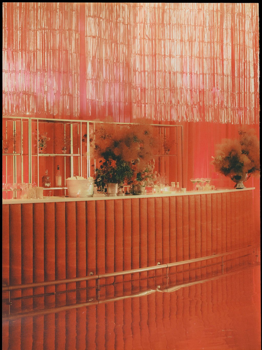 A bar or reception area with a coral-colored velvet front, decorative flowers, and glassware, against a coral wall with vertical metallic accents and a textured ceiling installation.