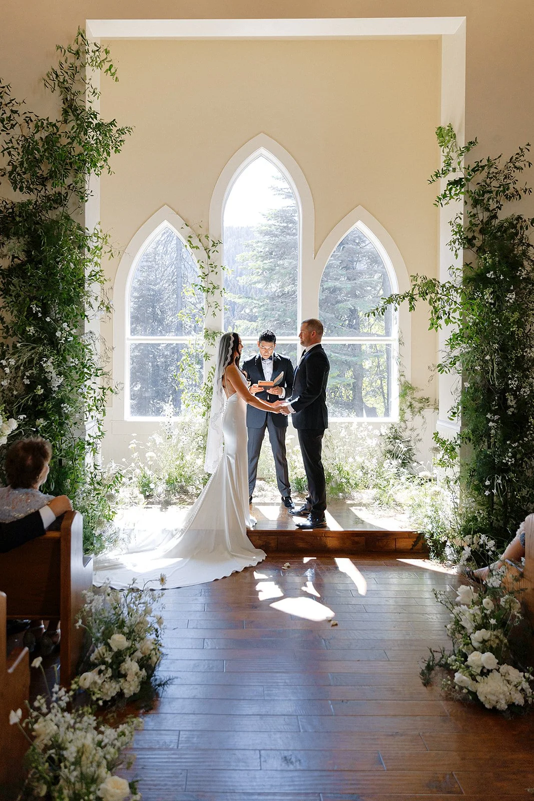 A couple gets married in a church with large arched windows, during daytime, with a officiant standing between them. The bride is wearing a white wedding dress and the groom is in a black suit. Guests are seated on benches, and there are white flower
